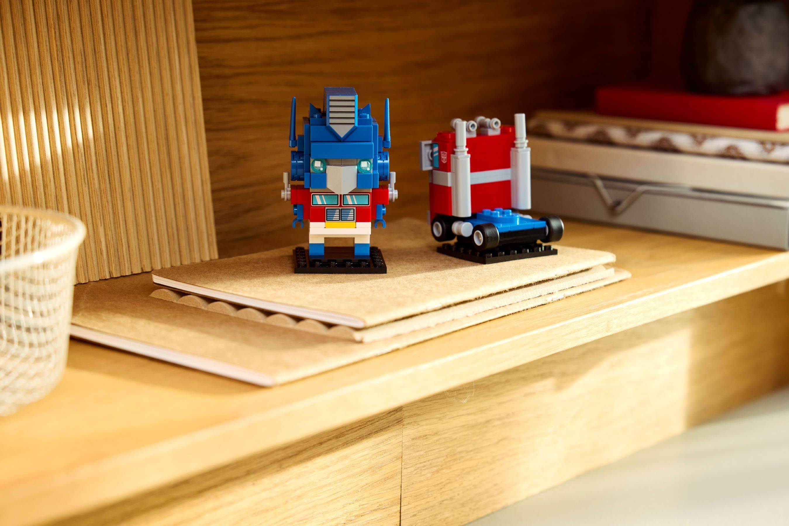 Zwei kleine LEGO-ähnliche Roboter auf einem Holzregal, einer blau und einer rot-weiß, auf einer beigefarbenen Matte ausgestellt.