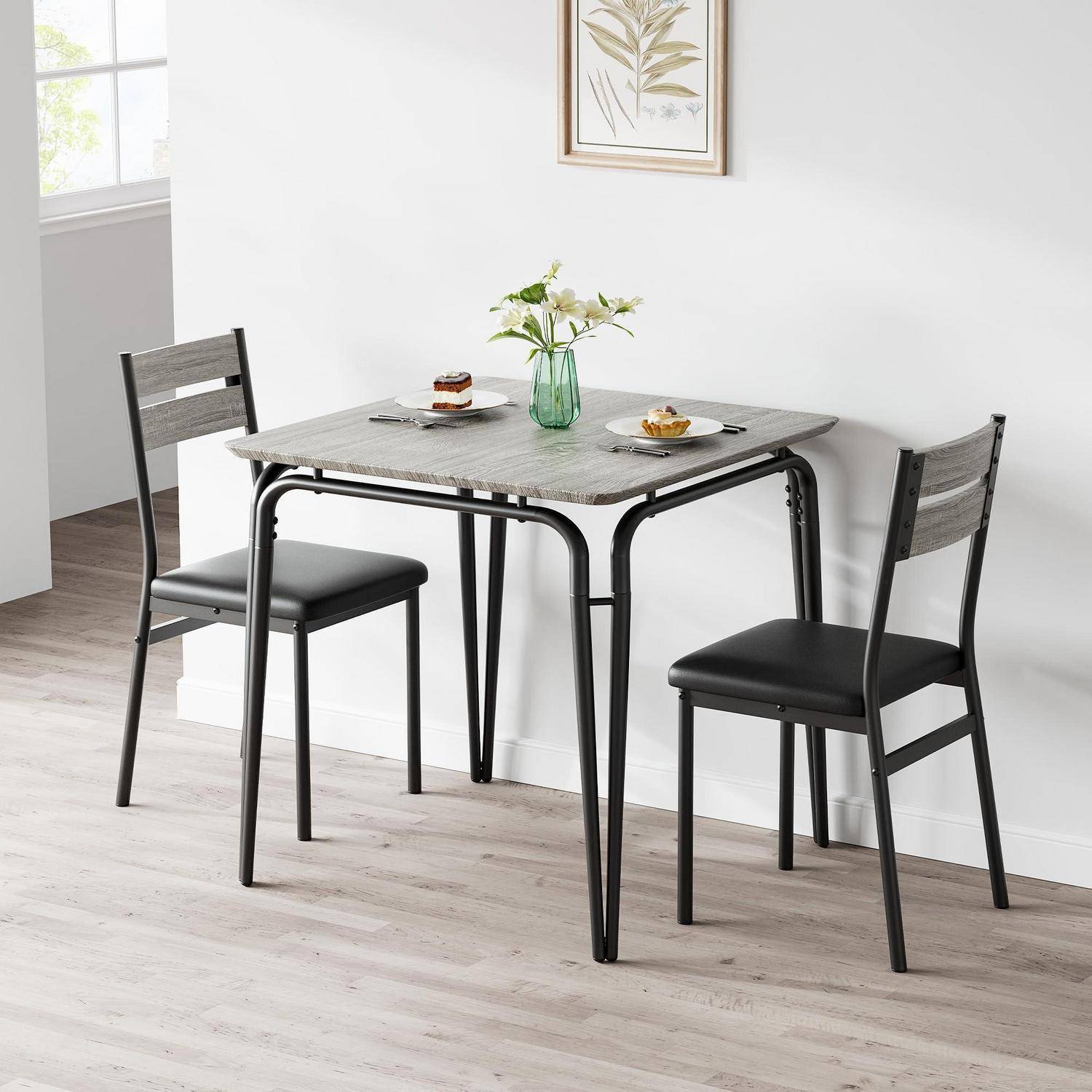 Kleiner Esstisch mit zwei schwarzen Stühlen in einem hellen modernen Raum, gedeckt mit Desserttellern und einer Blumenvase.