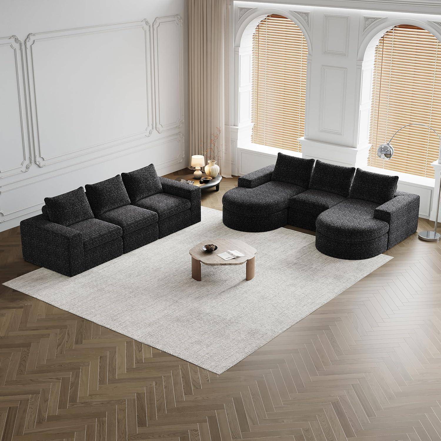 Modernes Wohnzimmer mit zwei schwarzen modularen Ecksofen, die sich gegenüberstehen, auf einem hellen Flächenteppich, mit einem niedrigen runden Couchtisch und großen Bogenfenstern mit Jalousien.