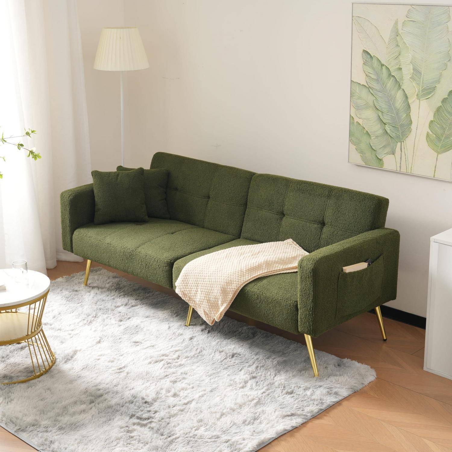 Grünes Samtstoffsofa mit goldfarbenen Beinen und einer Wurfdecke in einem hellen Wohnzimmer mit weißem Teppich, Stehleuchte und Blatt-Wandkunst.