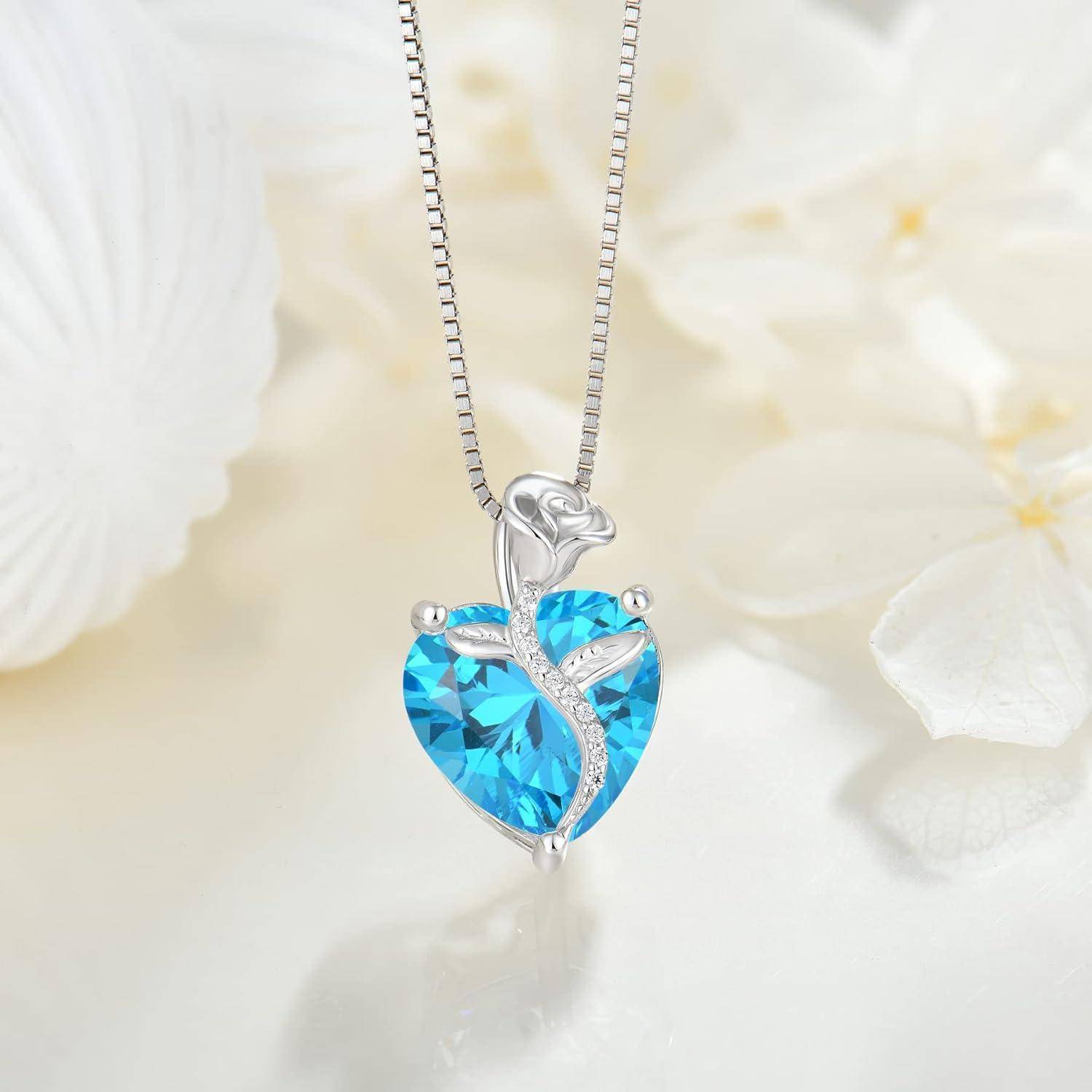 Blaue herzförmige Kristallanhängerkette mit silbernem Blattdesign an einer feinen Kette, vor einem weichen Blumenhintergrund dargestellt.