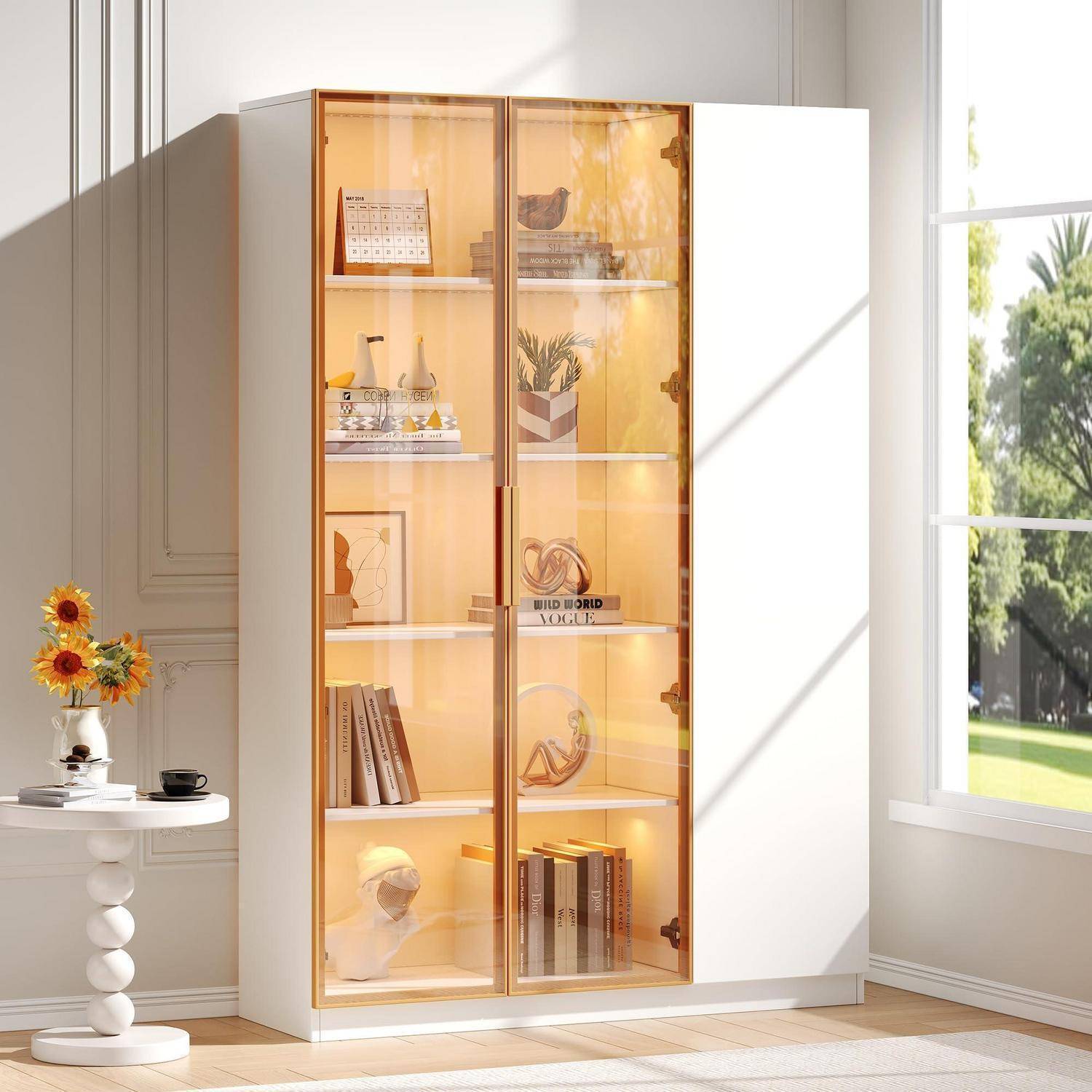 Weißer Schrank mit bernsteinfarbenen Glastüren und Regalen, die Bücher, Dekoration und Pflanzen in einem hellen Raum neben einem Fenster ausstellen.
