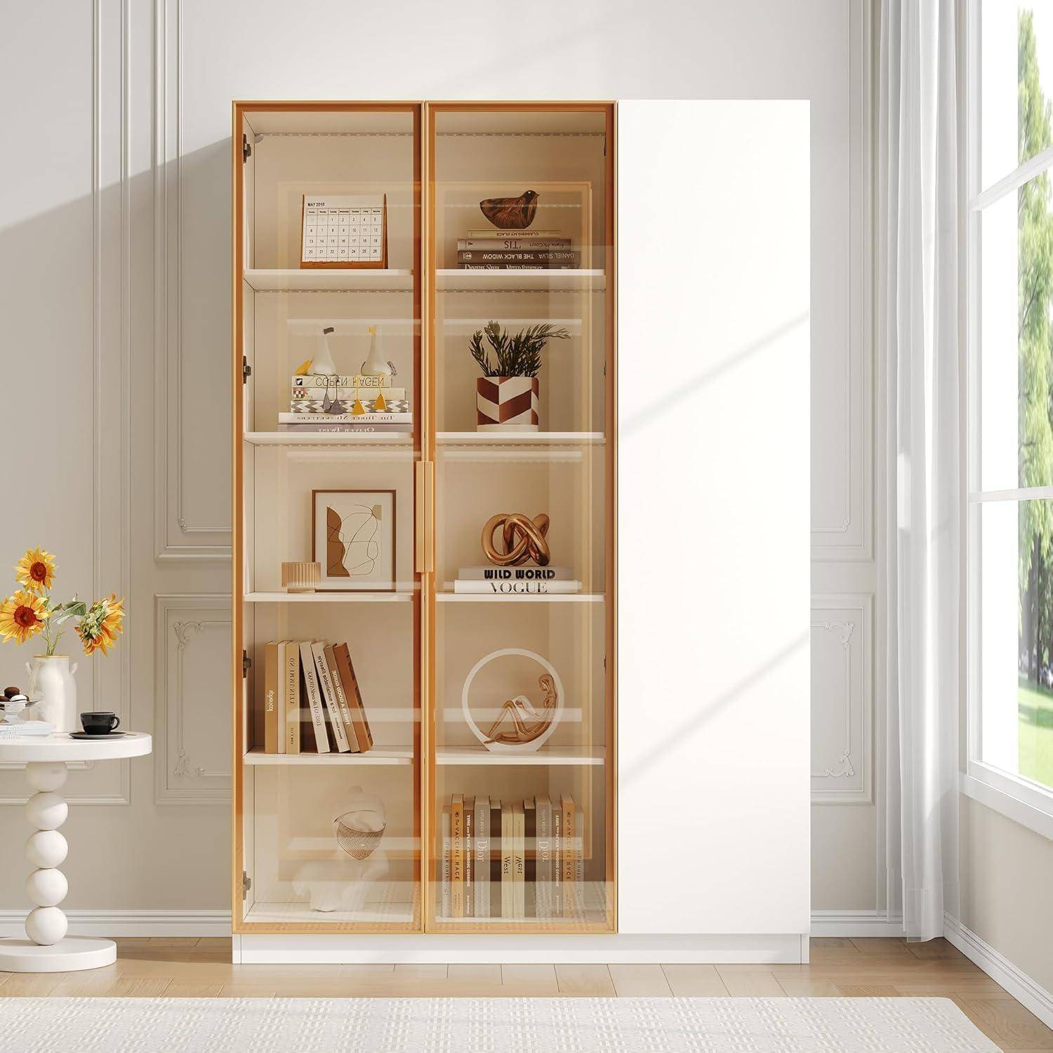Glassfront-Schrank mit Büchern und dekorativen Gegenständen auf Regalen in einem hellen Raum, neben einem schlichten weißen Paneel und Fenster.