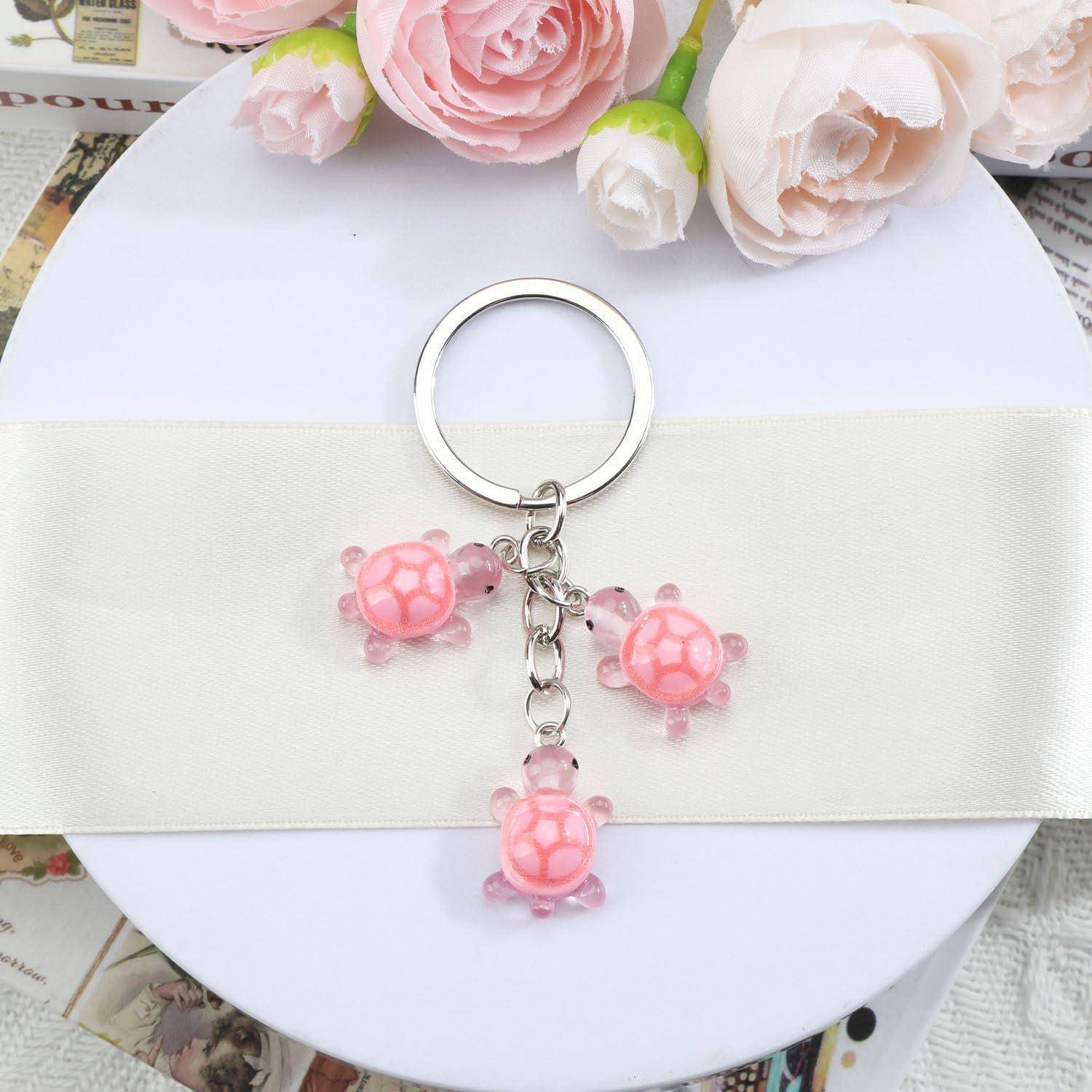 Silberner Schlüsselanhänger mit drei rosa Schildkröten-Anhängern, die von einem Ring an einem weißen Band über einer runden weißen Oberfläche hängen, mit Blumen oben.