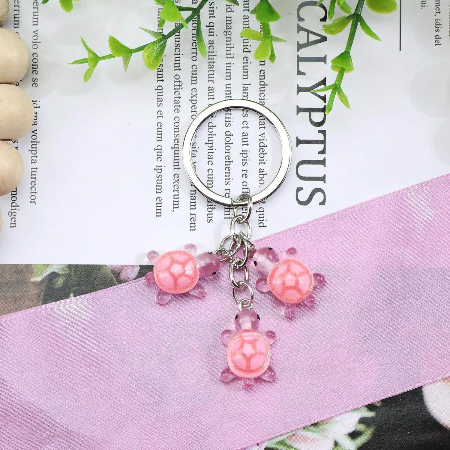 Rosa Schildkröten-Charme Schlüsselanhänger mit drei kleinen durchscheinenden Schildkröten-Perlen, die an einem Metallring befestigt sind, ruht auf einem rosa Band über bedrucktem Papier.