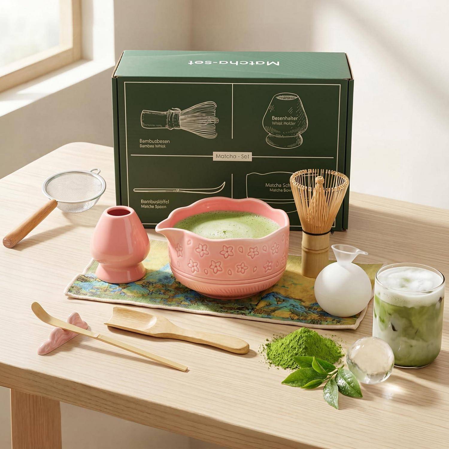 Matcha-Teeset auf einem Holztisch, bestehend aus einer rosa Whisk-Schale, einem Schneebesen, einem Löffel, einem Sieb und Tassen, mit einer grünen Produktbox im Hintergrund.