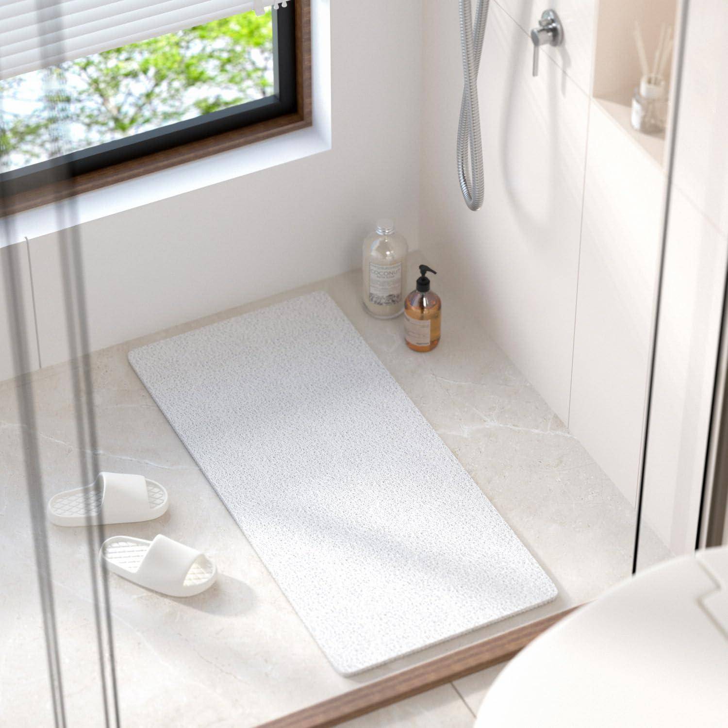Minimales Badezimmer mit weißer Badmatte, Pantoffeln, Shampooflaschen und Duschschlauch neben einem Fenster und einer gläsernen Duschtür.