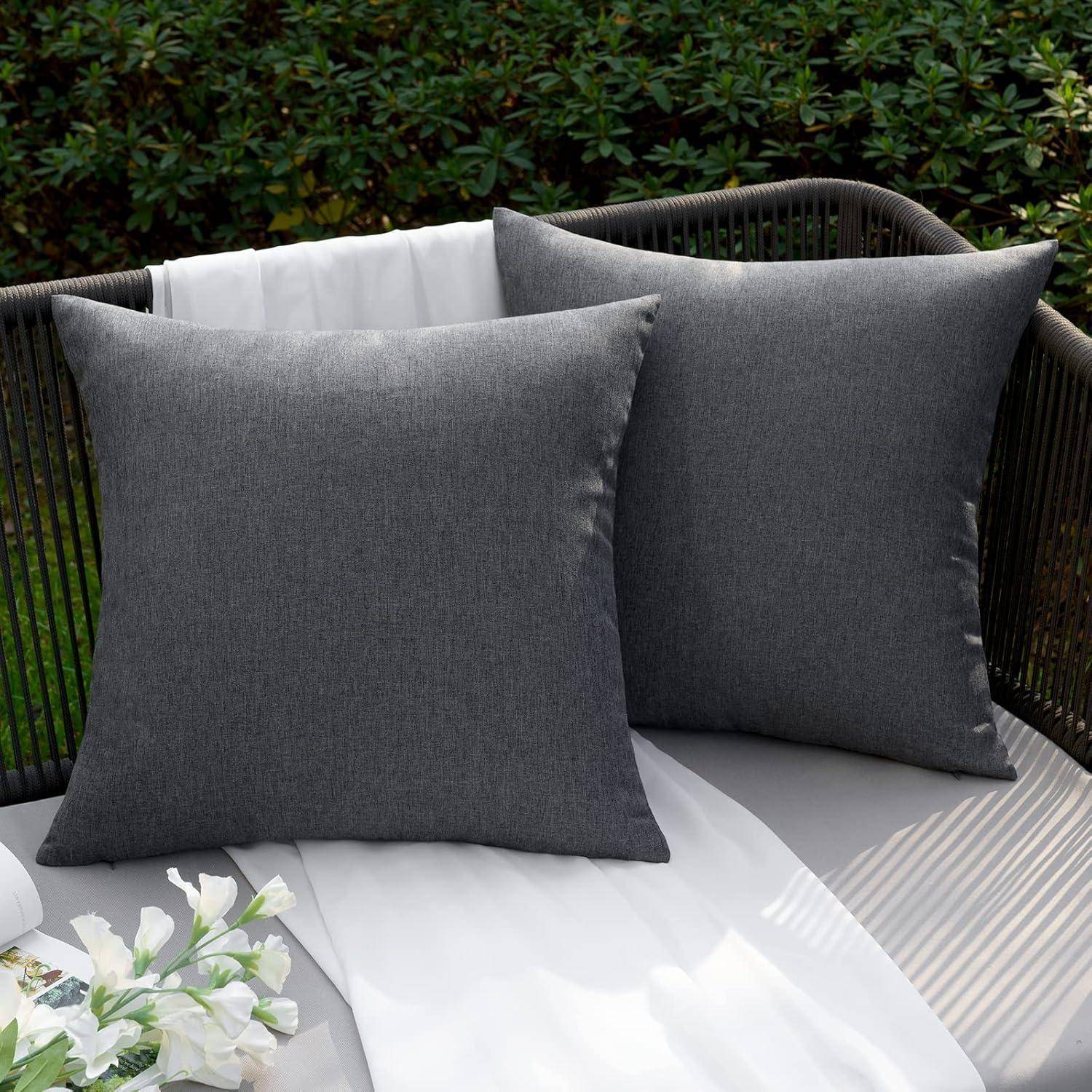 Zwei große graue Outdoor-Wurf-Kissen auf einem Korbsofa mit einer weißen Decke und kleinen weißen Blumen.