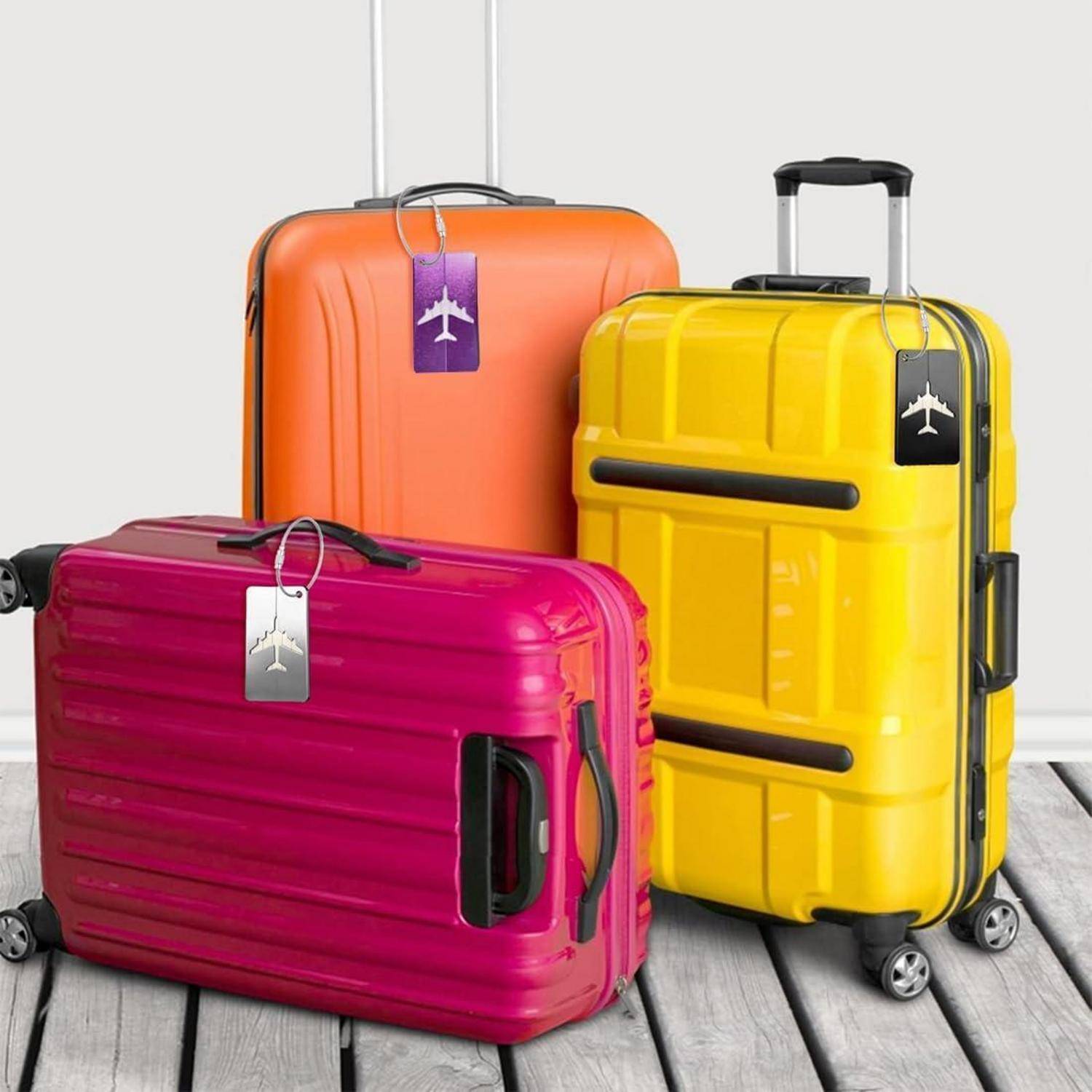 Drei bunte Hartschalenkoffer mit Gepäckanhängern: rosa vorne, orange dahinter und gelb aufrecht mit ausgeklapptem Griff auf einem Holzboden.