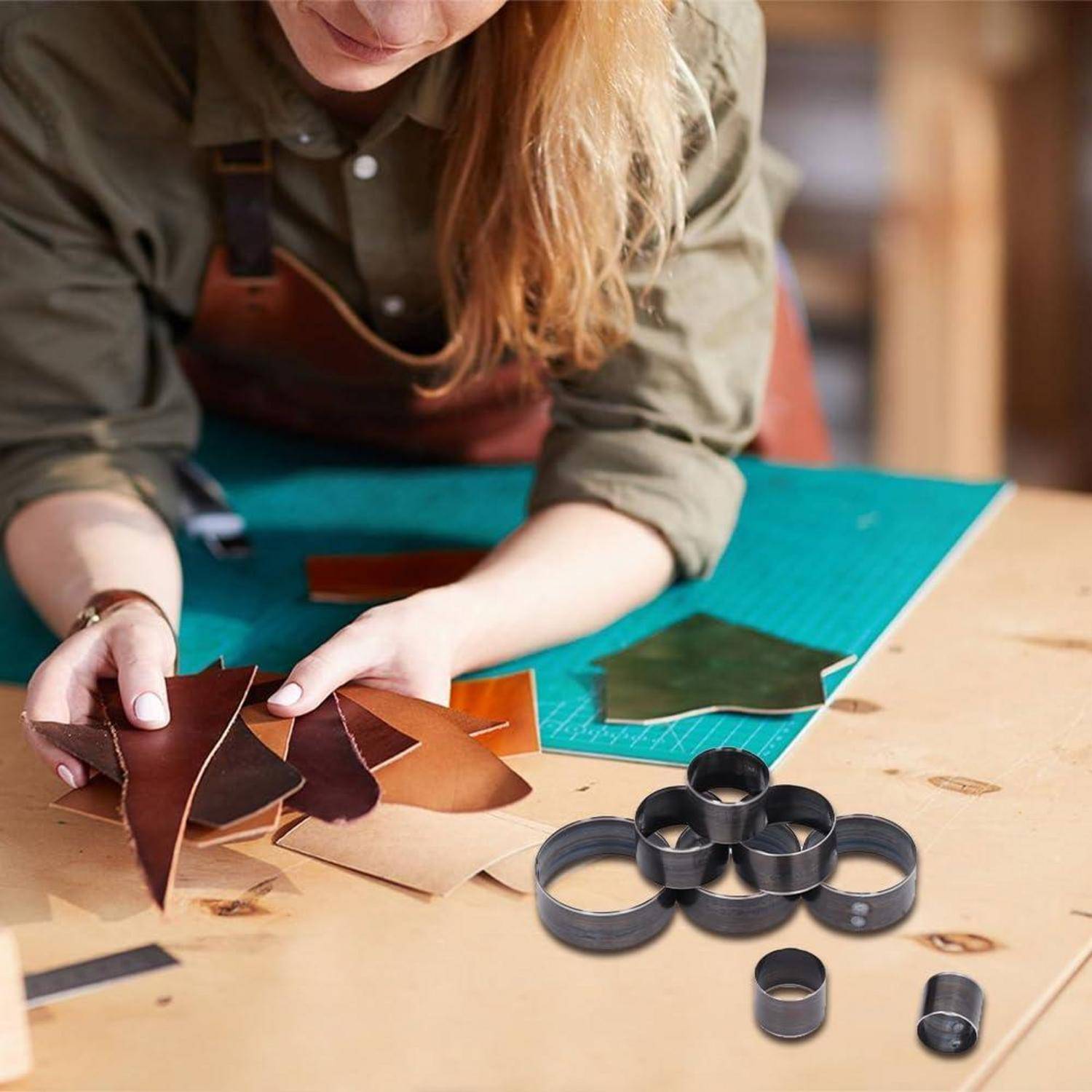 Person, die mit Lederstücken auf einer Schneidematte arbeitet, geschnittene Formen und Metallringwerkzeuge auf einem Handwerkstisch anordnet.
