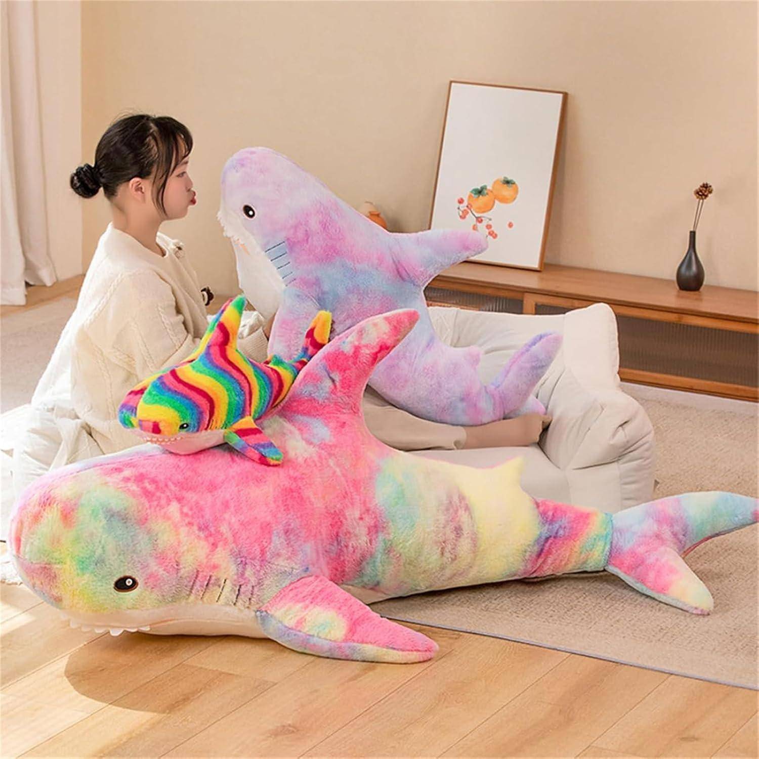 Frau sitzt auf einem Sofa mit drei großen regenbogenfarbigen Batik-Plüsch-Haifisch-Spielzeugen in einem Wohnzimmer.