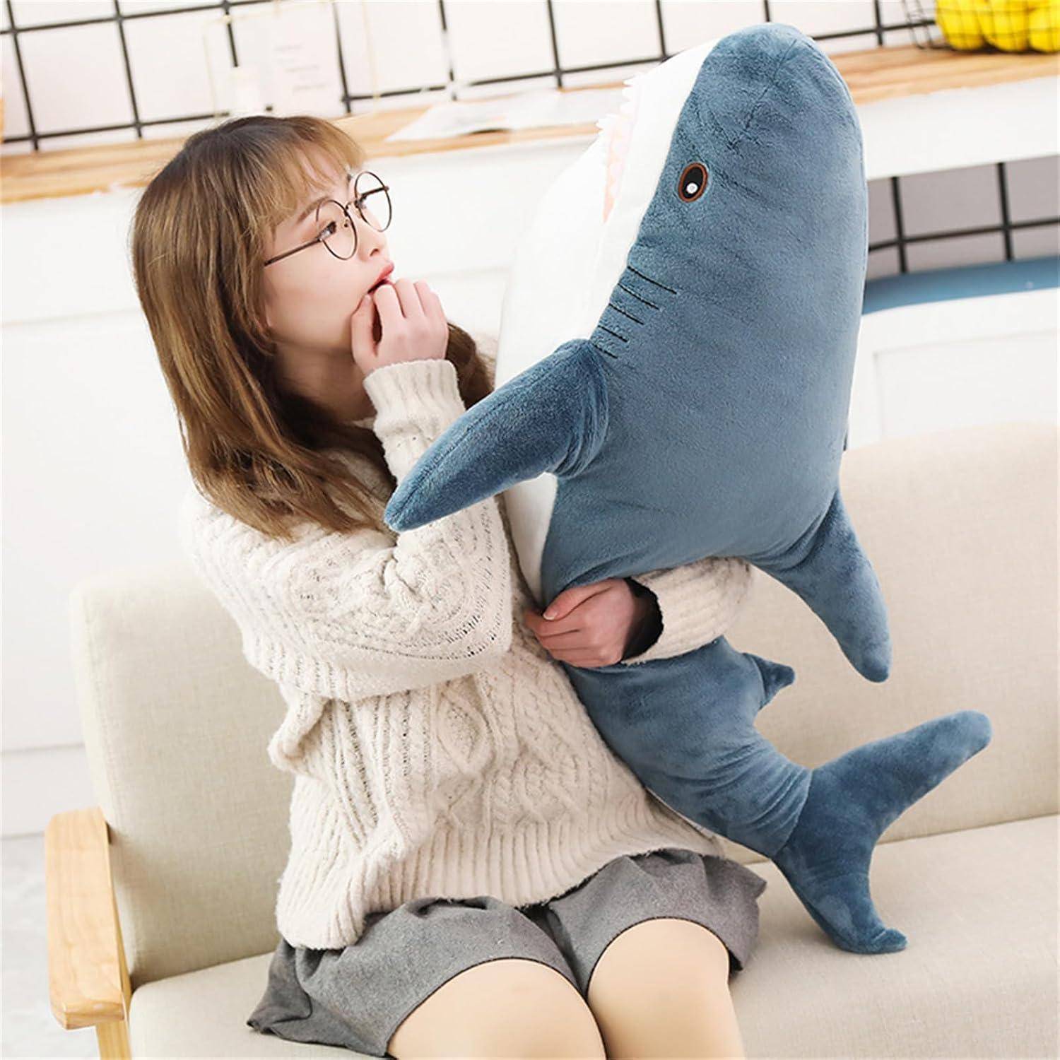 Ein junges Mädchen mit Brille sitzt auf einem Sofa und umarmt einen großen blauen Plüschhai, während es ihn überrascht anschaut.