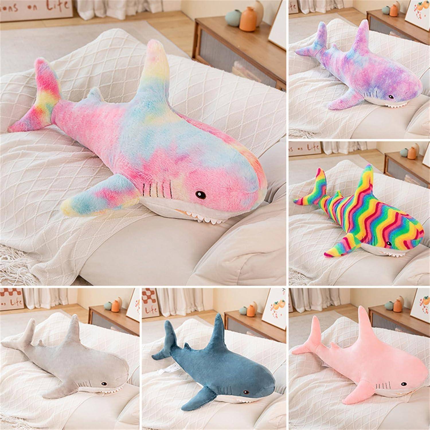 Collage aus kuscheligen Haifisch-Kuscheltieren auf einem Bett in Pastelltönen, Regenbogen-, Weiß-, Blau- und Rosatönen.