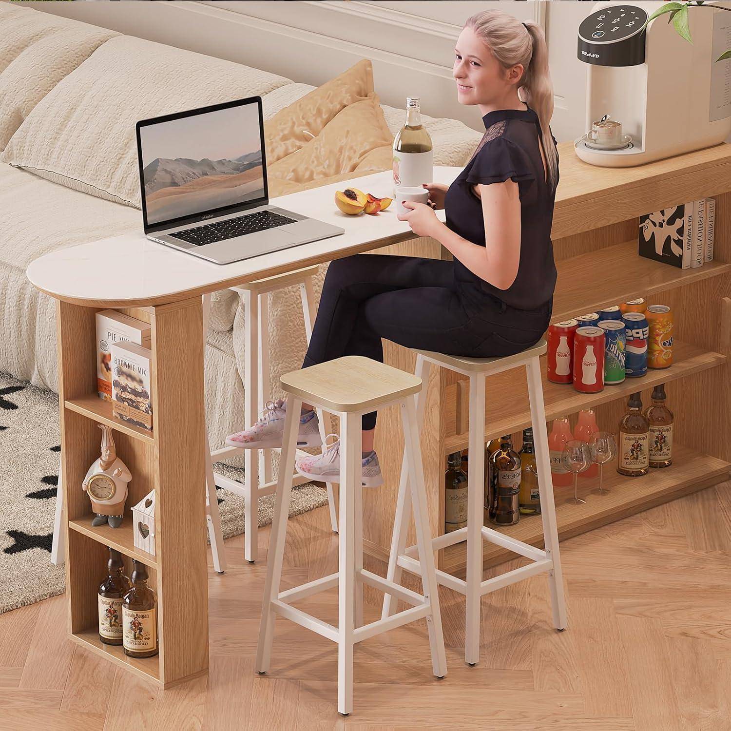 Frau, die an einem kompakten Nachttisch mit Laptop und Getränk sitzt, nutzt einen kleinen Heimarbeitsplatz, der mit Regalen für Snacks, Flaschen und Bücher integriert ist.