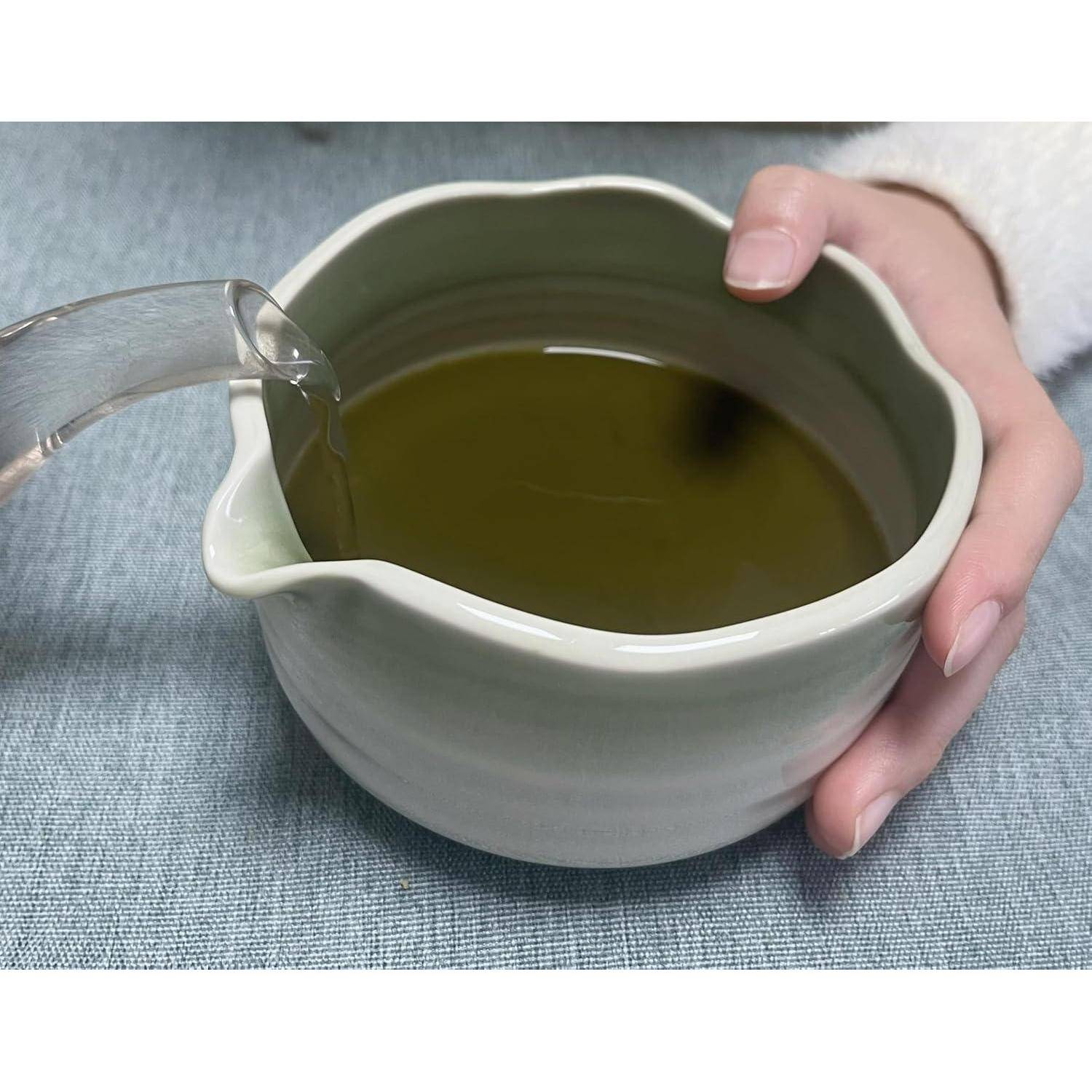 Eine Hand gießt Wasser in eine Keramikschale mit grünem Tee auf einer Tischdecke.