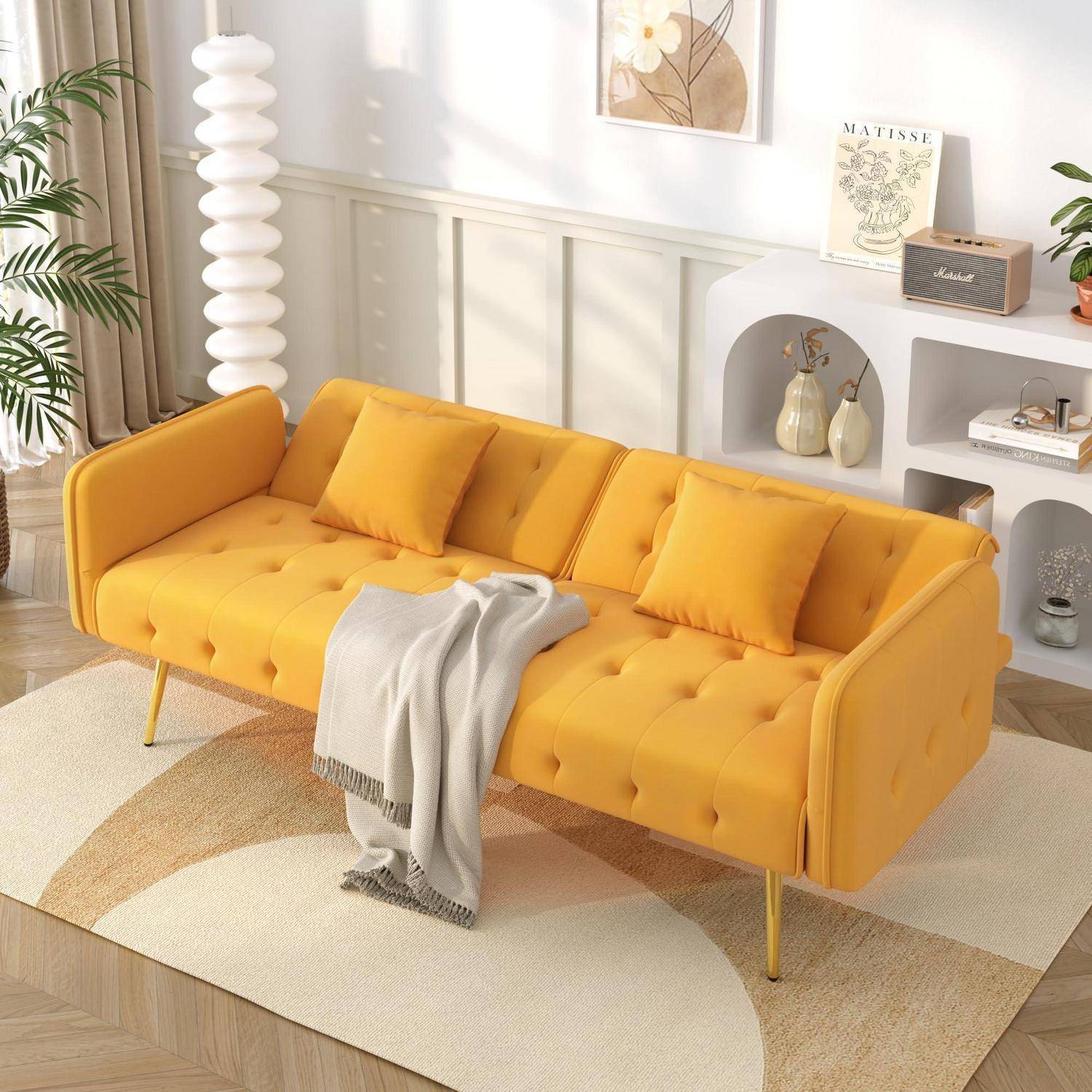 Modernes Wohnzimmer mit einem senfgelben, gestepptem Sofa, drei Kissen und einer Wurfdecke, eingerichtet mit weißen Regalen und Dekoration in weichem Tageslicht.