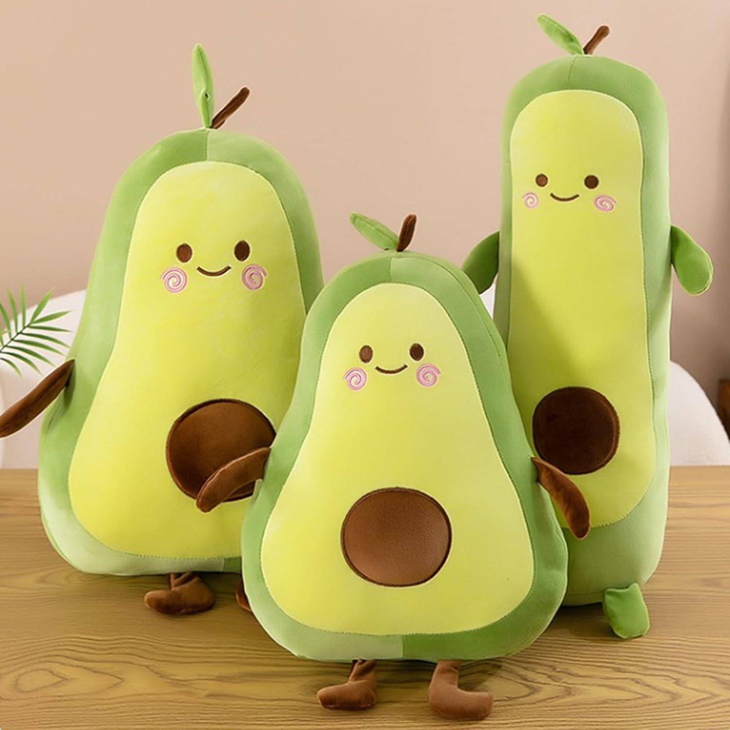 Drei plüschige Avocado-Spielzeuge mit lachenden Gesichtern und braunen Kernen sitzen auf einer Holzoberfläche vor einem beigen Hintergrund.