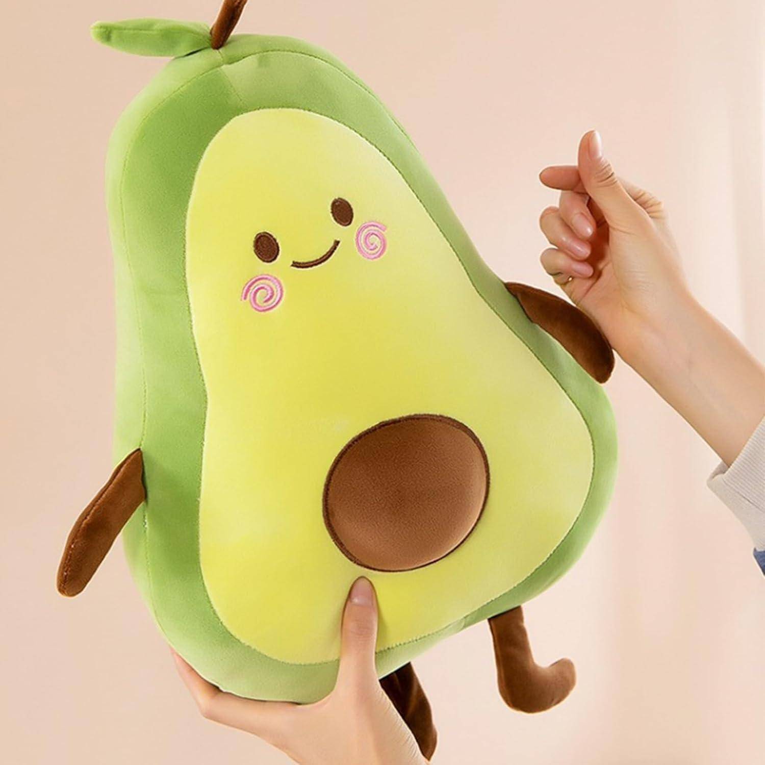 Kuscheliges Avocado-Spielzeug mit einem lächelnden Gesicht und braunem Kern, von zwei Händen gegen einen einfachen Hintergrund gehalten.