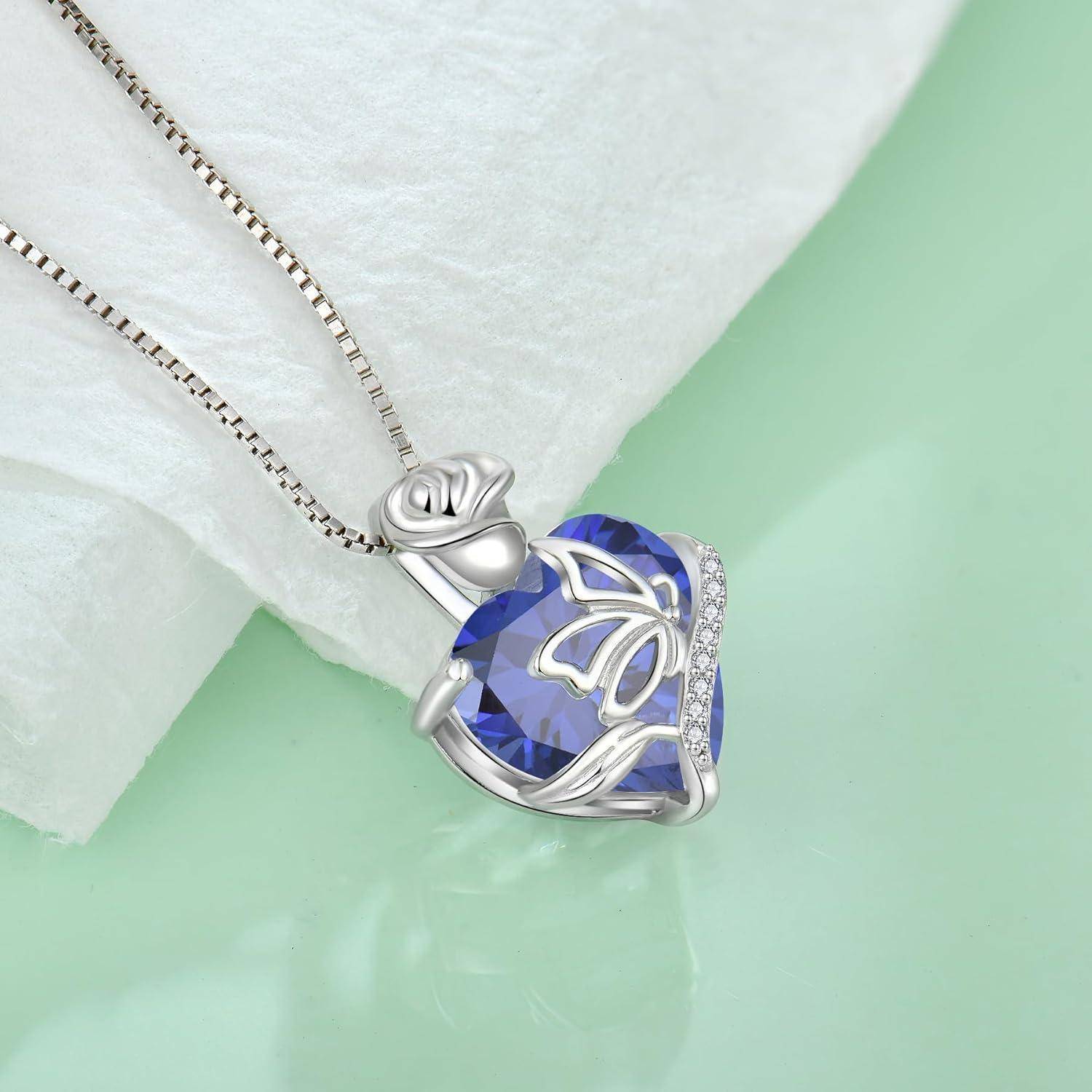 Silberanhänger-Halskette mit einem blauen, facettierten, herzförmigen Edelstein und Schmetterlingsdesign an einer Kette, ausgestellt auf weißem Stoff.