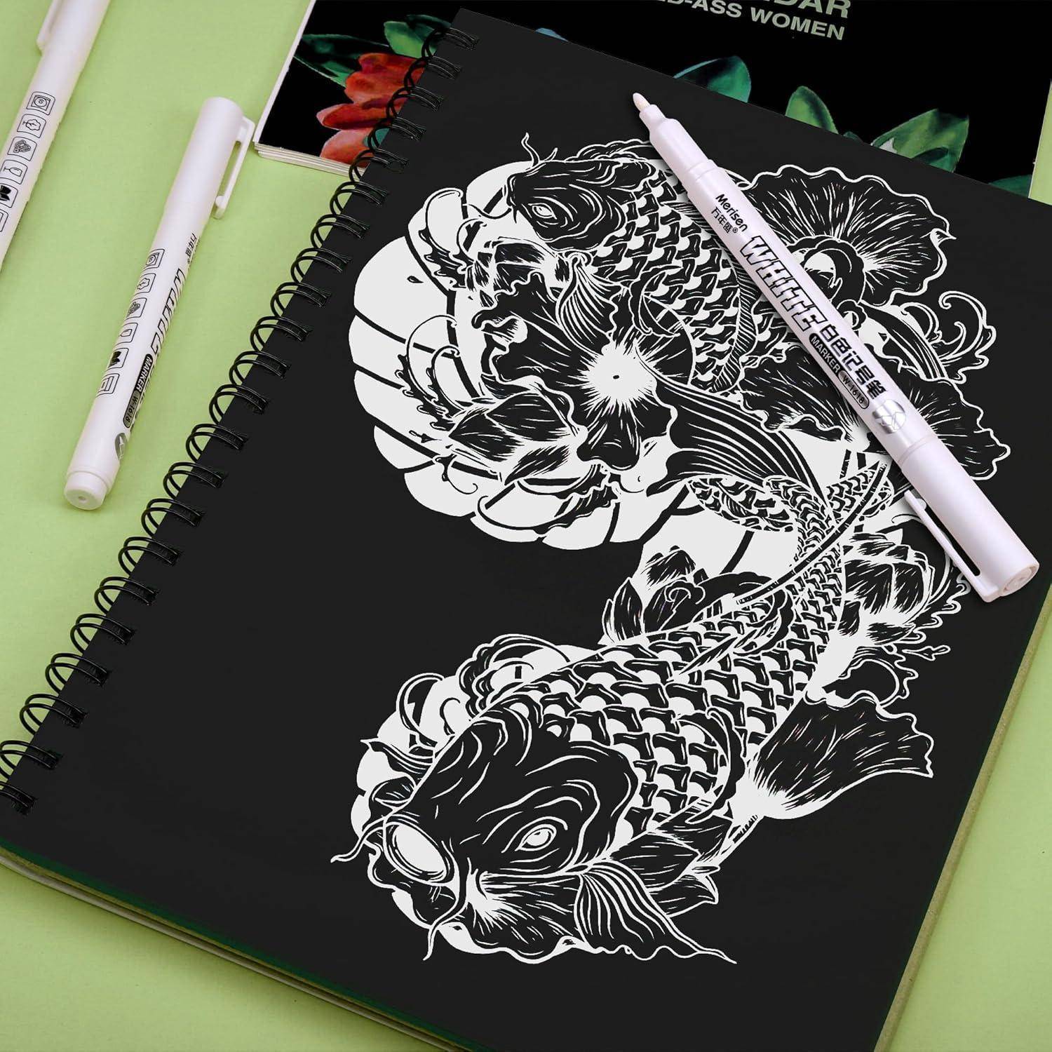 Schwarzes spiralgebundenes Notizbuch mit weißen Koi-Fischen und floralen Linienzeichnungen, überkreuzt von zwei weißen Markern, auf einer hellgrünen Oberfläche.