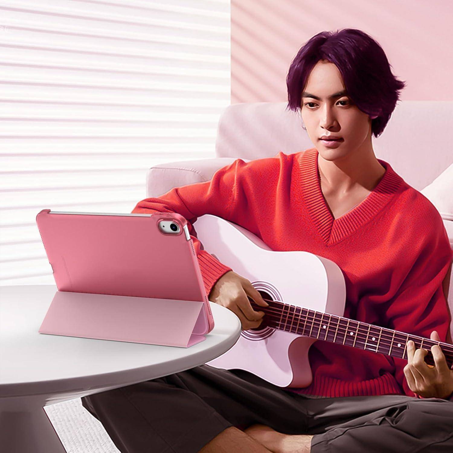 Ein junger Mann in einem roten Pullover sitzt mit einer weißen Gitarre neben einem rosa Tablet auf einem Tisch und scheint zu Hause Musik zu üben.