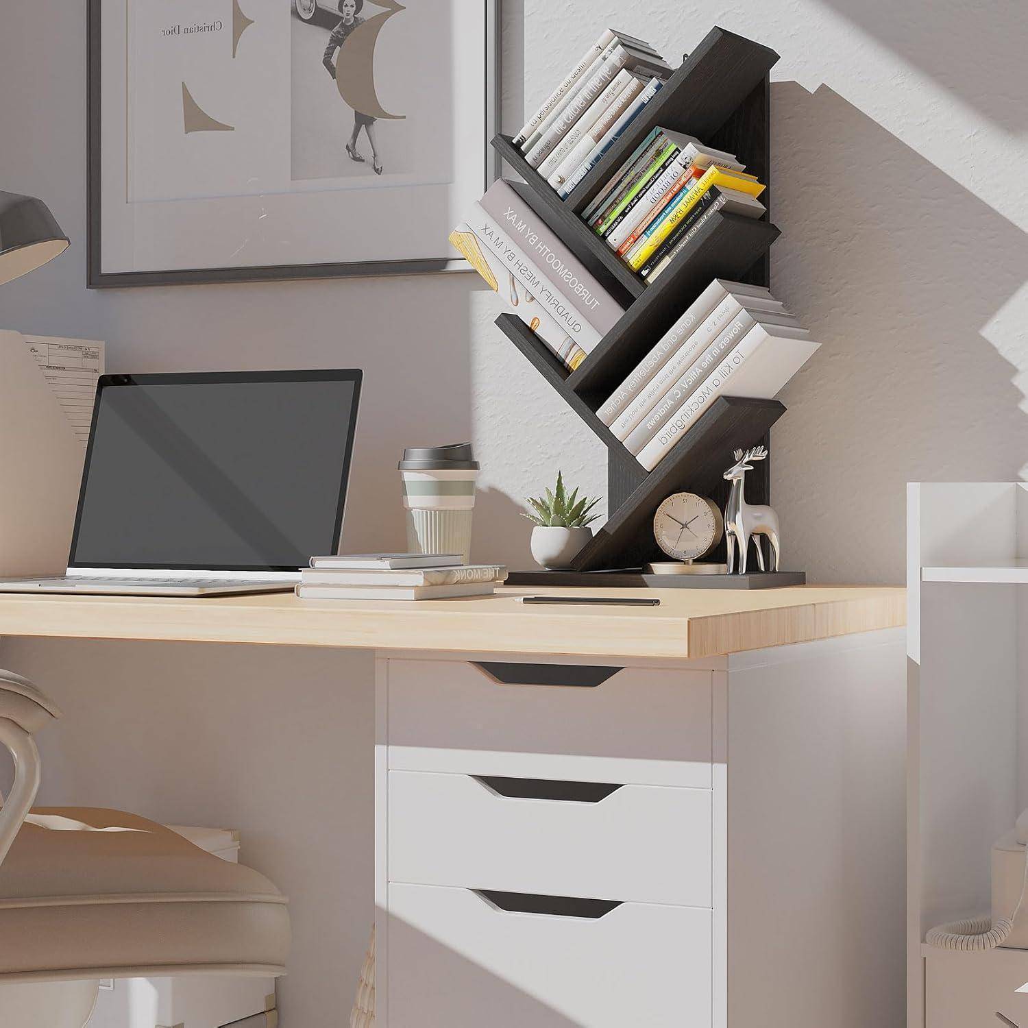 Moderner Schreibtisch im Homeoffice mit Laptop, gestapelten Büchern, Kaffeetasse, einer kleinen Pflanze, Uhr und einem schräg angebrachten Wandregal über einer weißen Schubladeneinheit.