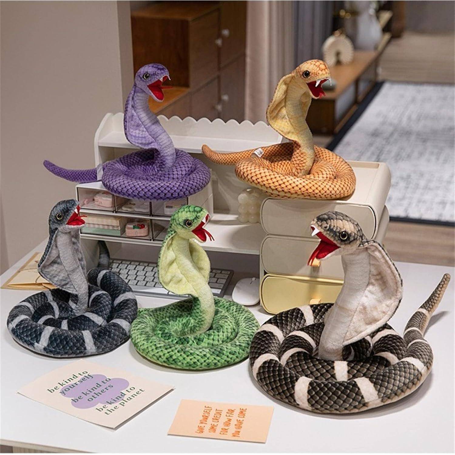 Fünf gestrickte Schlangen-Plüschtiere in verschiedenen Farben sind auf einem Schreibtisch rund um eine Miniatur-Computeranlage angeordnet.