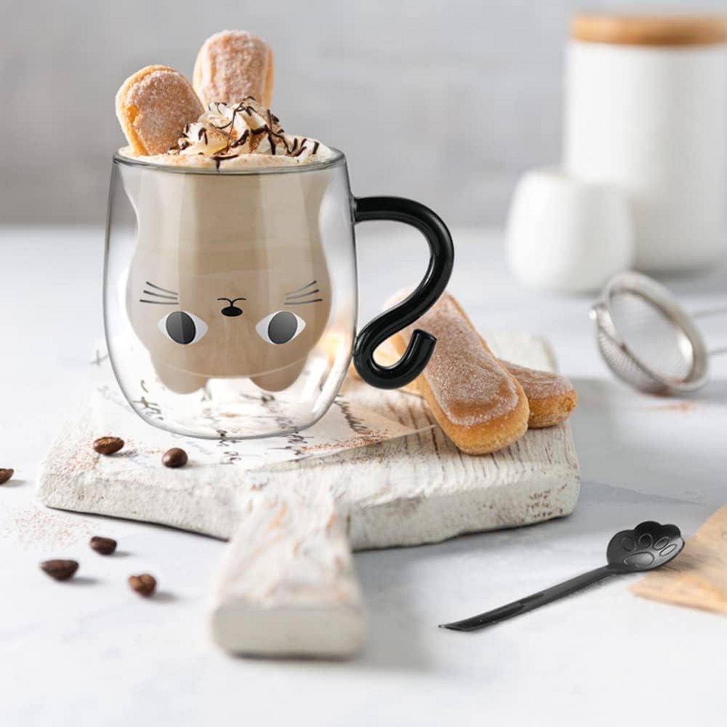 Katzengesicht-Glastasse mit Schlagsahne und Ladyfinger-Keksen, stilisiert auf einem rustikalen Brett mit verstreuten Kaffeebohnen und Löffel.