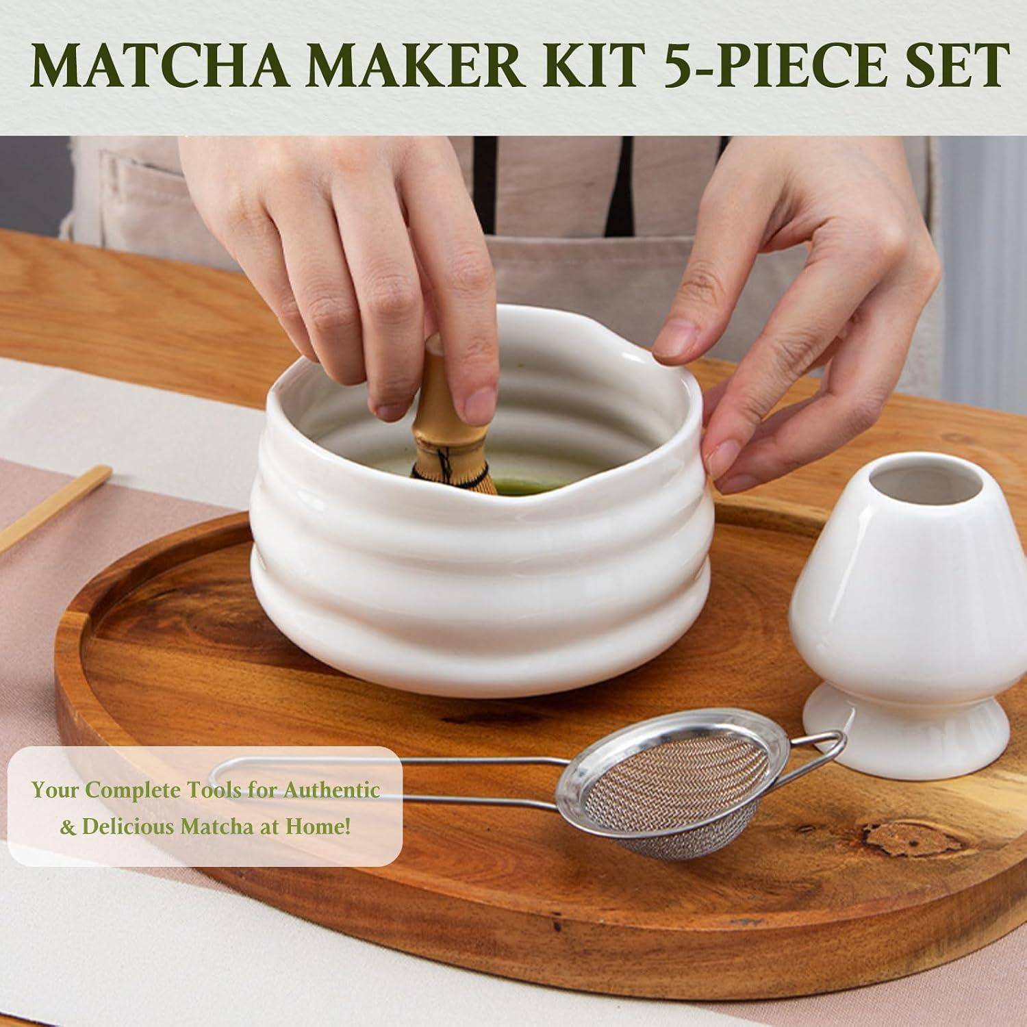 Produktbild eines „MATCHA-MAKER-SETS MIT 5 TEILEN", das Hände zeigt, die Matcha in einer weißen Schüssel mit einem Sieb und einem Ständer auf einem Holztablett schlagen.