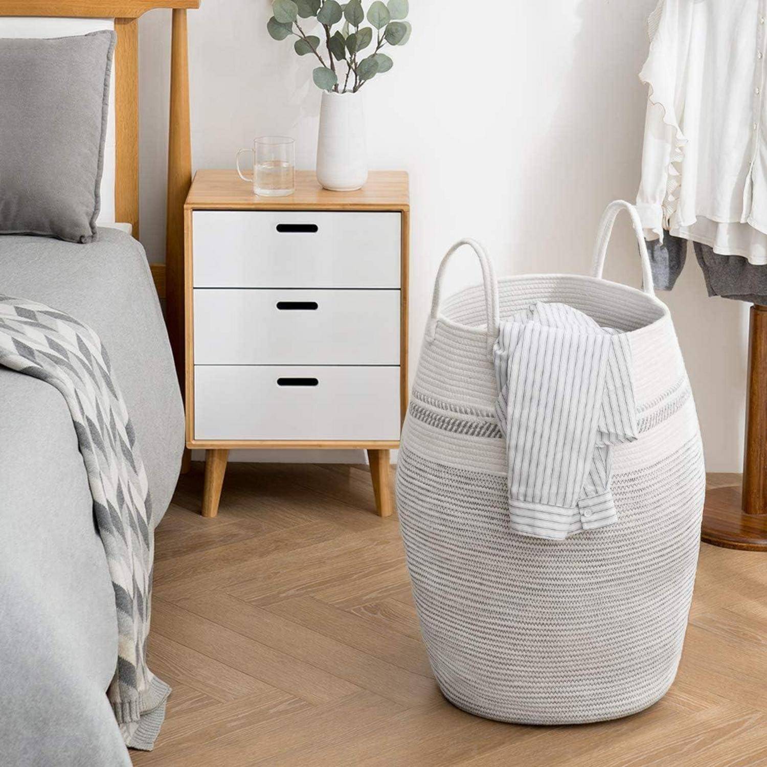 Weiß geflochtener Wäschekorb mit Griffen und gestreiftem Handtuch in einem Schlafzimmer neben einem Bett und einem Holznachttisch mit Schubladen und einer Vase.