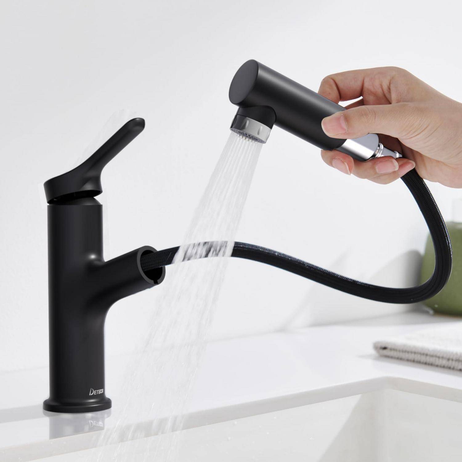 Hand, die eine schwarze ausziehbare Sprühdüse hält, die an einem mattschwarz Badezimmerhahn befestigt ist, und Wasser in ein Waschbecken leitet.