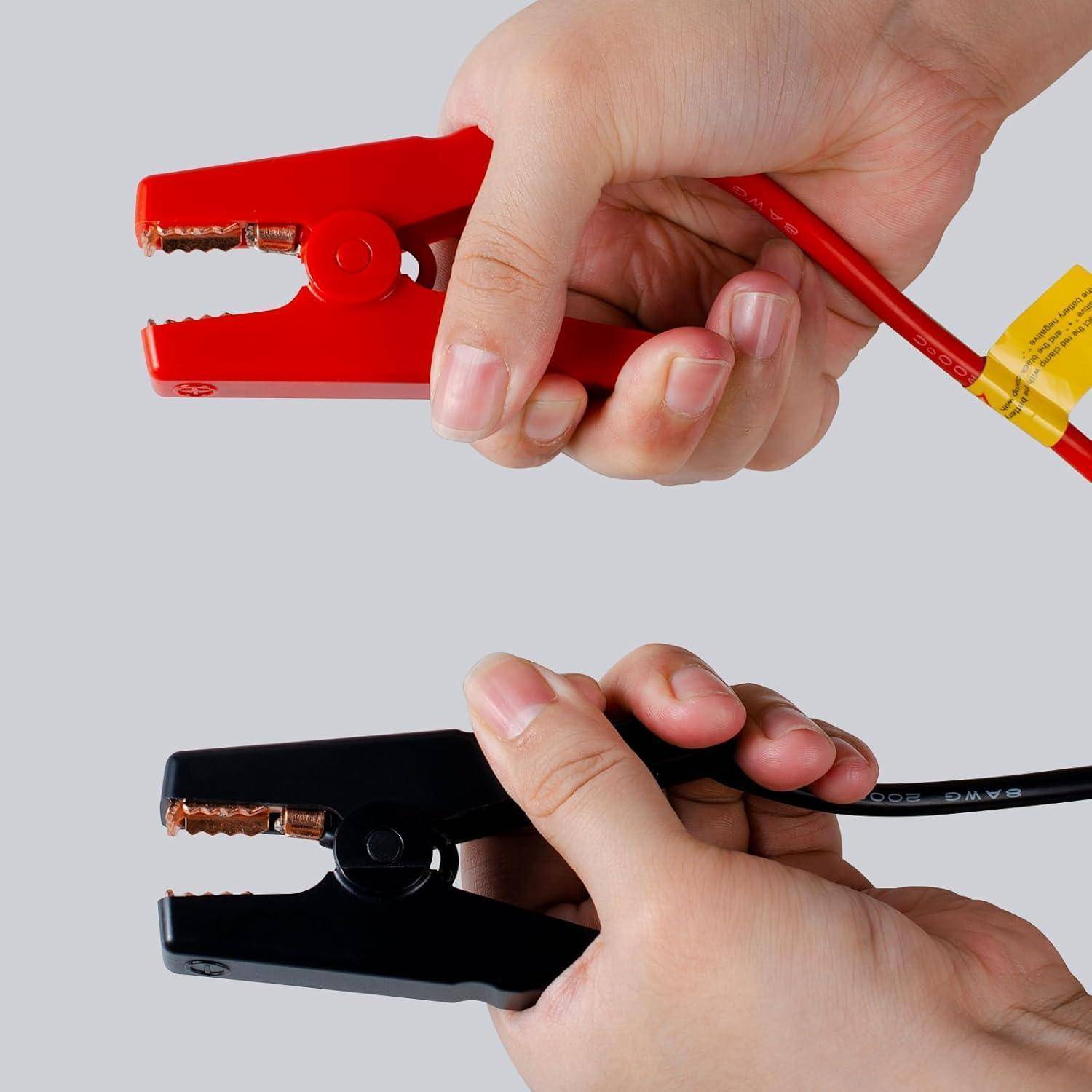 Zwei Hände halten rote und schwarze Autobatterie-Überbrückungsklemmen mit angeschlossenen Kabeln vor einem einfachen grauen Hintergrund.