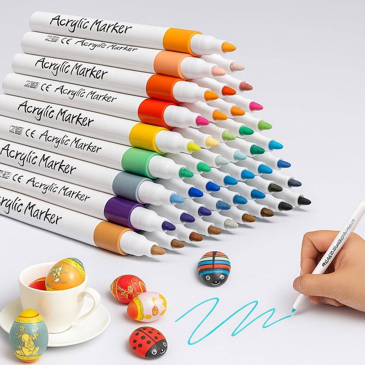 Sortierte Acrylmarker in Reihen angeordnet, mit einer Hand, die ein blaues Gekritzel zeichnet, und bemalten dekorierten Eiern sowie einer Teetasse in der Ecke.