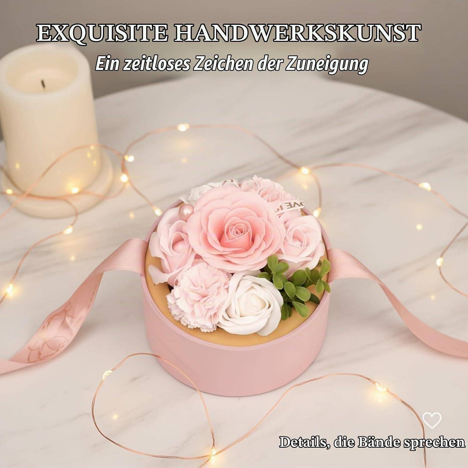 Rosa Blumenbox mit künstlichen Rosen und Schleife auf einer Marmoroberfläche, mit Kerze und Feenlichtern; Text lautet „EXQUISITE HANDWERKSKUNST