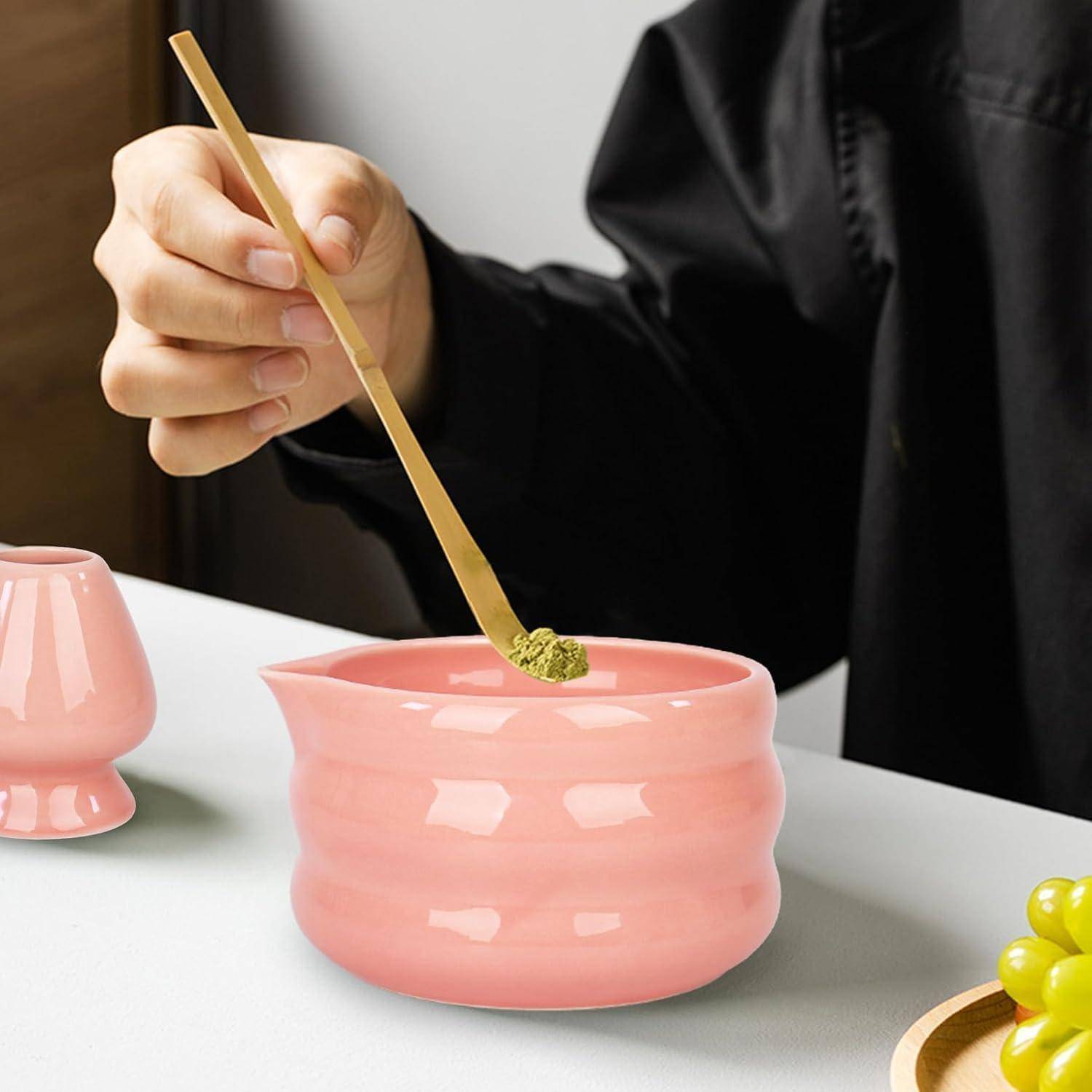 Hand, die einen Bambus-Matcha-Besen über eine rosa Keramikschale mit grünem Teepulver hält, mit einer passenden rosa Tasse und Trauben auf einem Tablett in der Nähe.