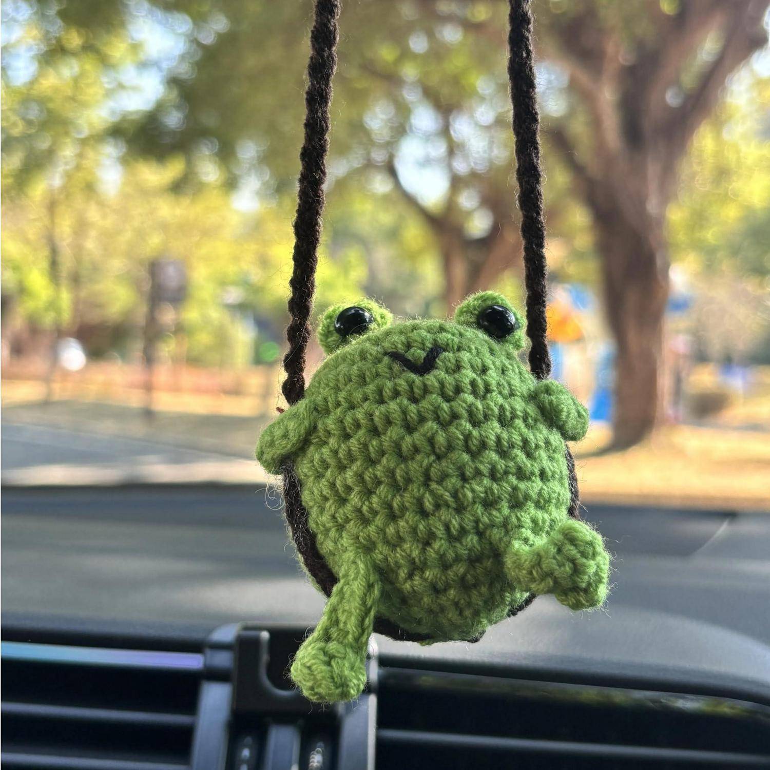 Ein kleines grünes gehäkeltes Frosch-Ornament hängt an einem Autoinnenrückspiegel, mit einer verschwommenen Außenstraße und Bäumen, die durch die Windschutzscheibe sichtbar sind.