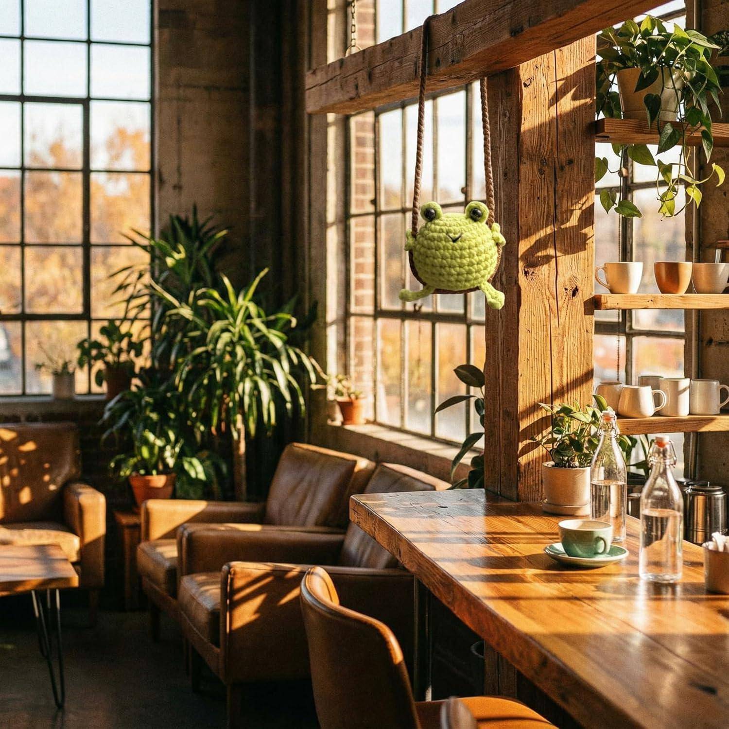 Sonniges, gemütliches Café-Interieur mit Lederstühlen, Holztischen und Regalen voller Pflanzen und Tassen, plus einer kleinen grünen Frosch-Dekoration, die am Fenster hängt.