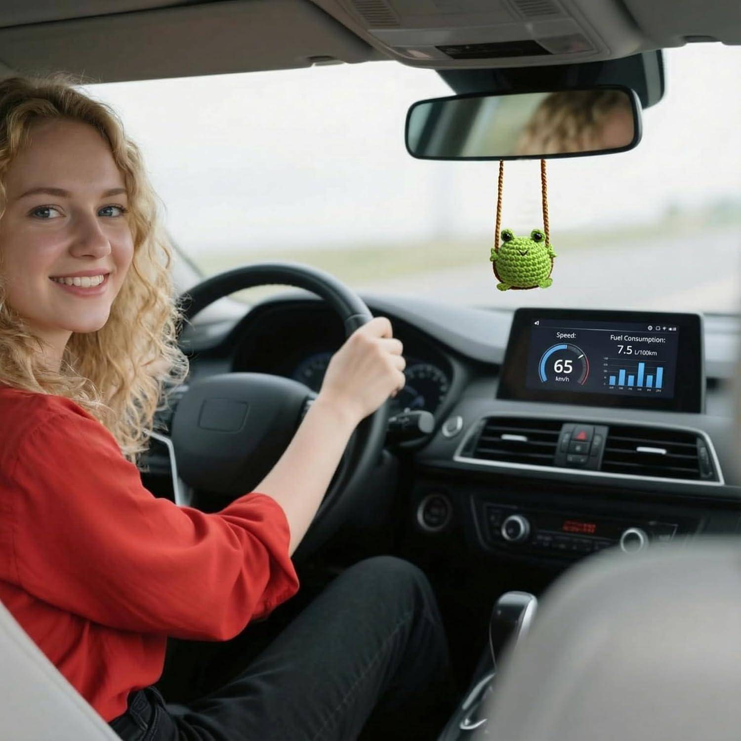 Lächelnde Frau, die ein Auto fährt, mit einem Armaturenbrett-Display, das eine Geschwindigkeit von 65 und einen Kraftstoffverbrauch von 7,5 l/100 km anzeigt, und einem Frosch-Ornament, das am Spiegel hängt.