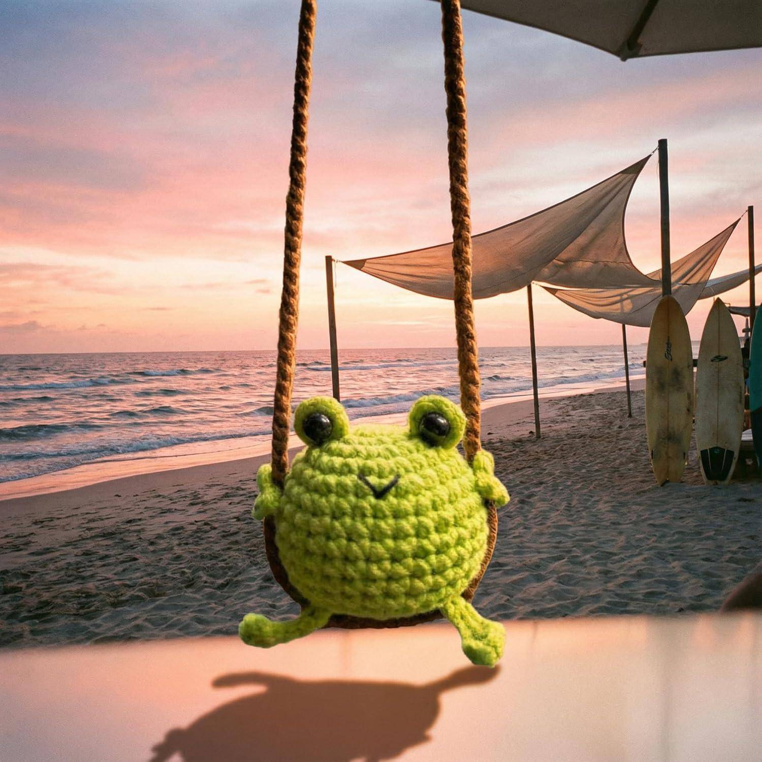 Eine gehäkelte grüne Frosch sitzt auf einer Schaukel am Strand bei Sonnenuntergang, mit Ozeanwellen, Sonnensegeln und Surfbrettern im Hintergrund.