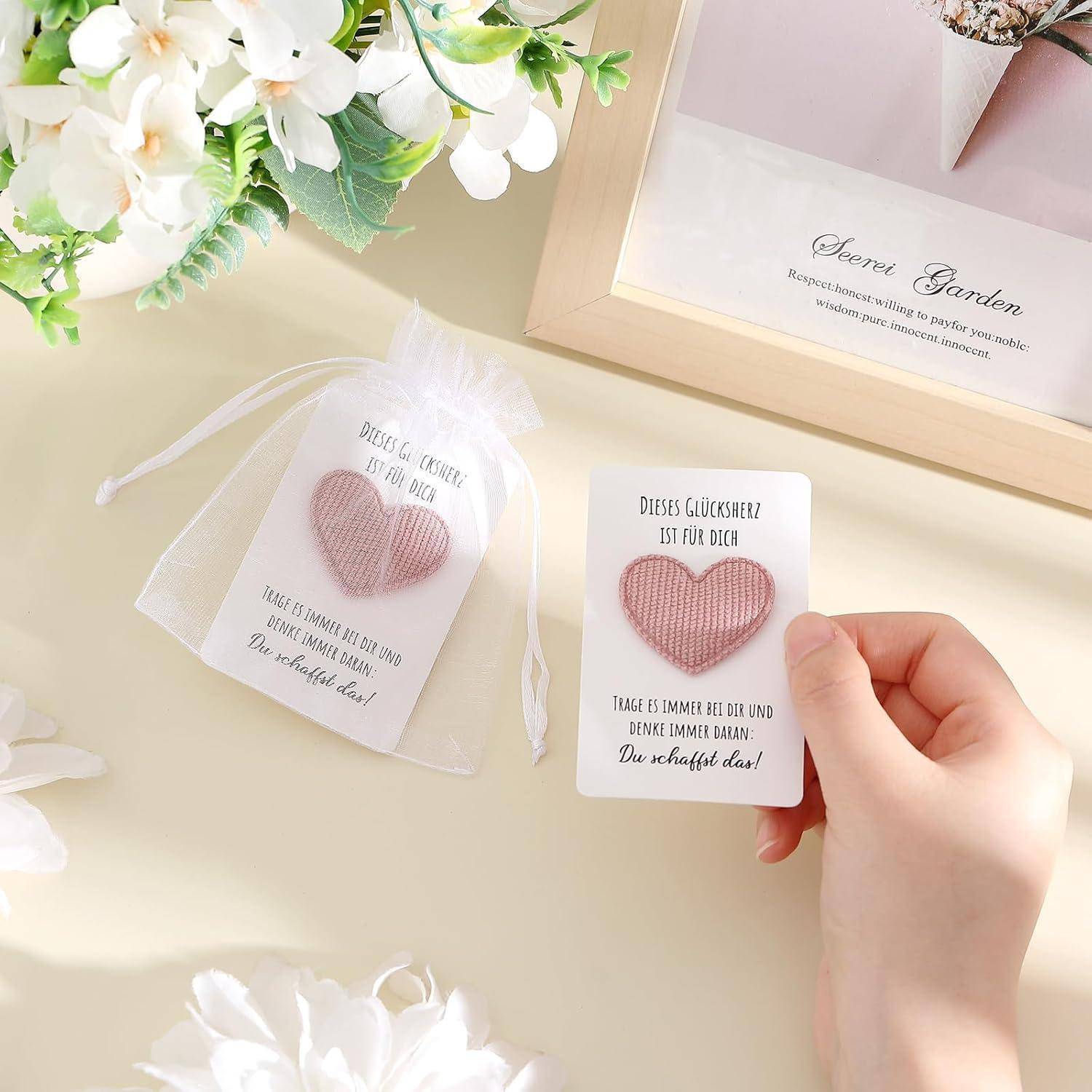 Eine Hand hält eine Geschenkkarte mit einem rosa Herz und deutschem Text, neben einer passenden Karte in einem durchsichtigen Beutel und einer 