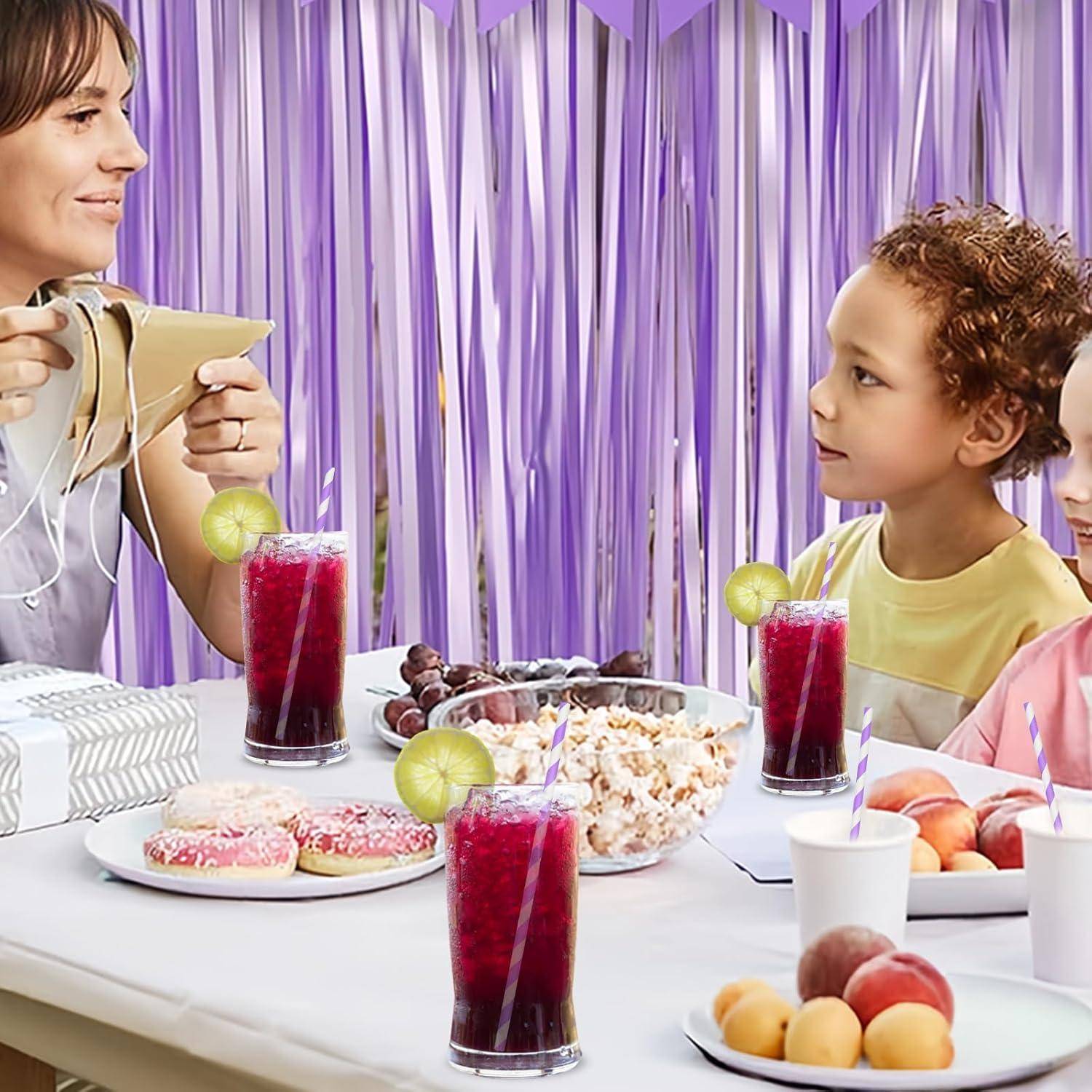 Frau, die ein Getränk an einem Kinderfest-Tisch ausschenkt, mit Saft, Donuts, Popcorn und Obst vor sitzenden Kindern mit lila Girlanden hinter ihnen.