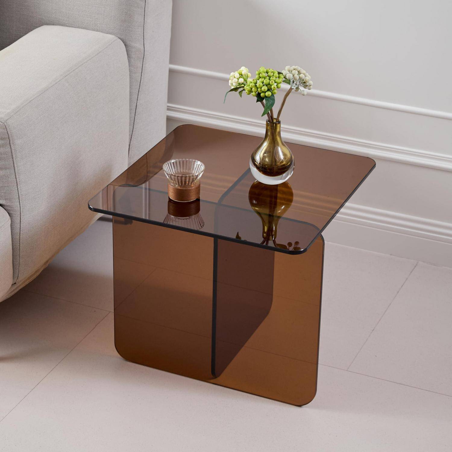 Moderner Beistelltisch aus braunem getöntem Glas neben einem beigen Sofa, mit einer kleinen Vase mit Blumen und einer Kerze oben drauf.