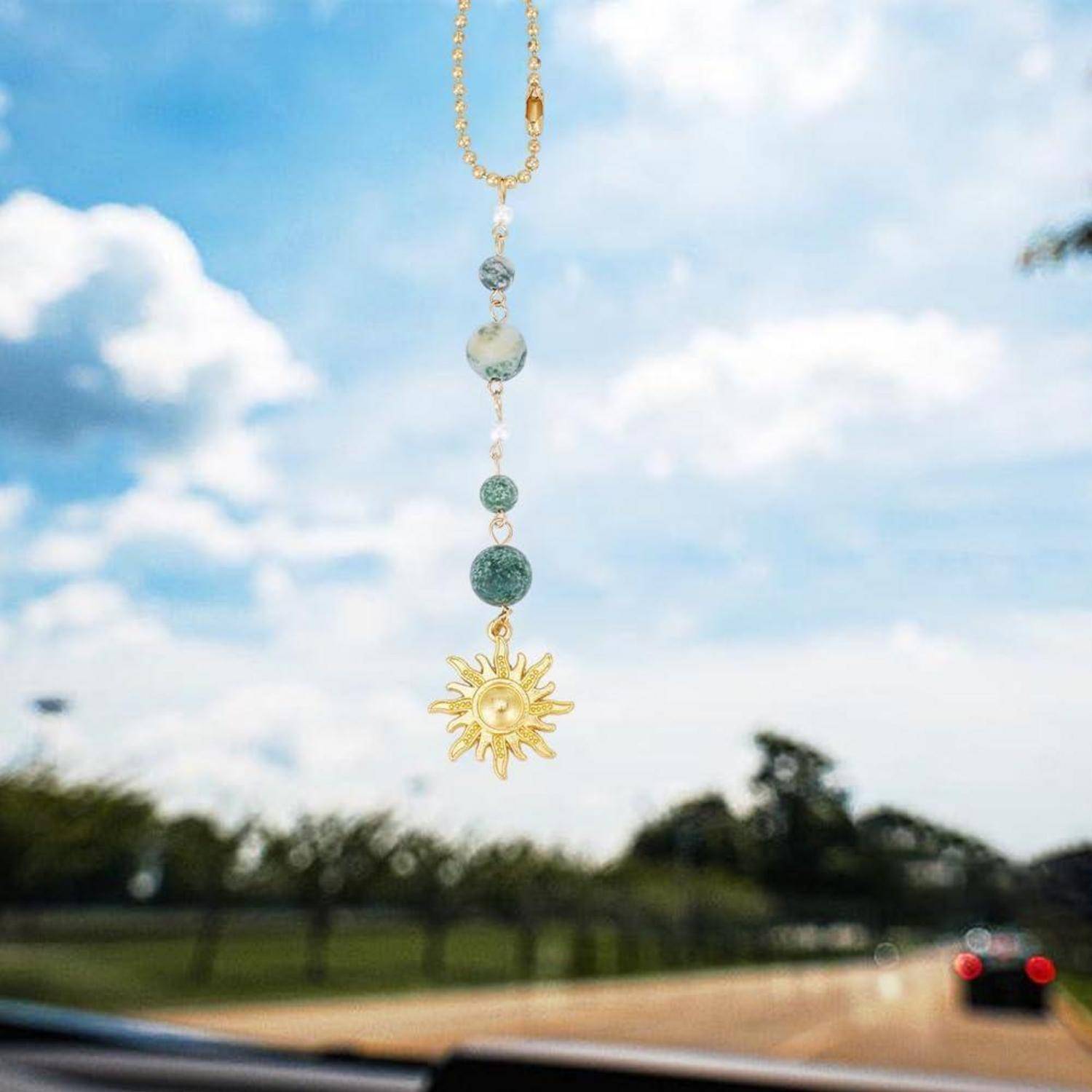 Eine goldene Halskette mit hellblauen Perlen und einem Sonnenanhänger hängt vor einer Windschutzscheibe eines Autos auf einer sonnigen Straße.