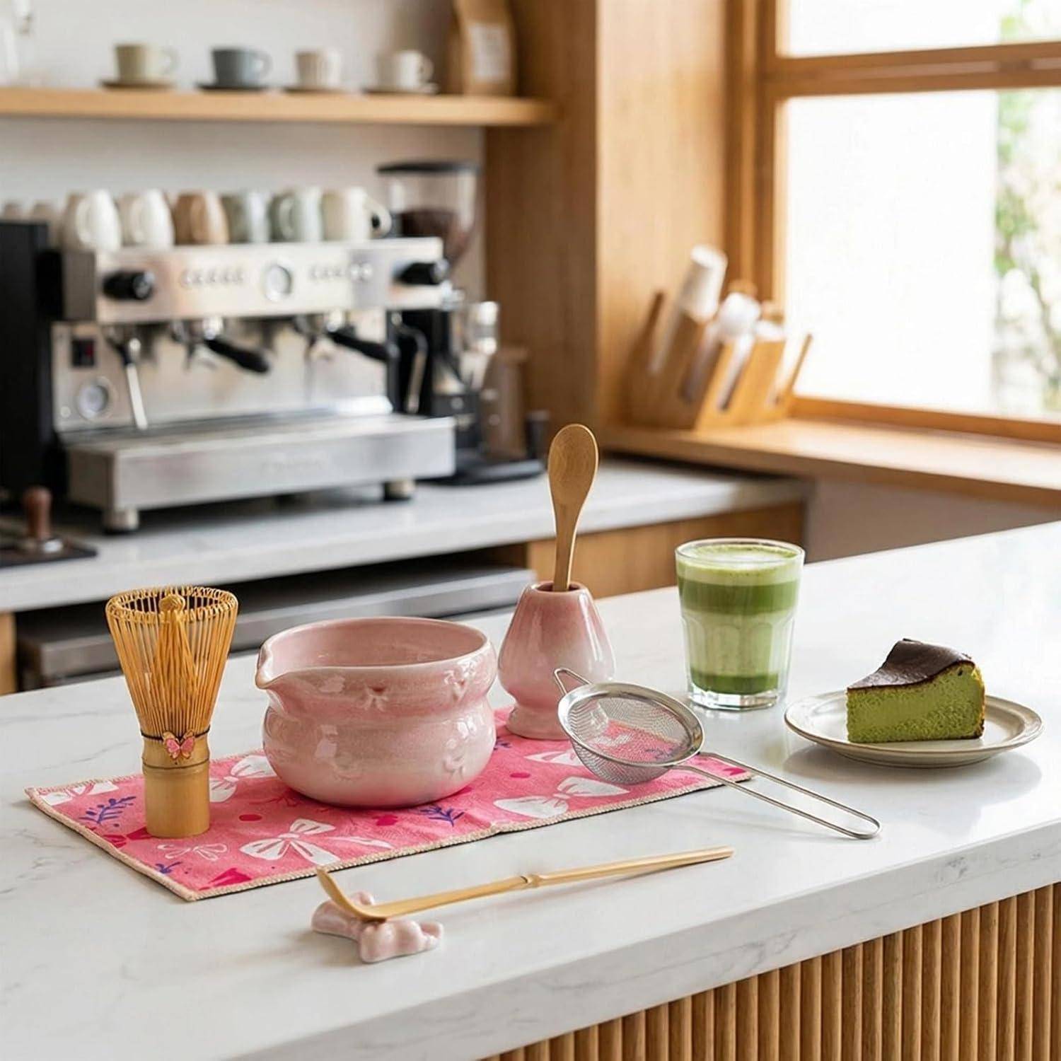 Ein Matcha-Tee-Set auf einer Küchentheke mit einem Schneebesen, einer Schale, einem Sieb, einer Tasse grünem Matcha und einem Stück Kuchen neben einer Espressomaschine und einem Fenster.