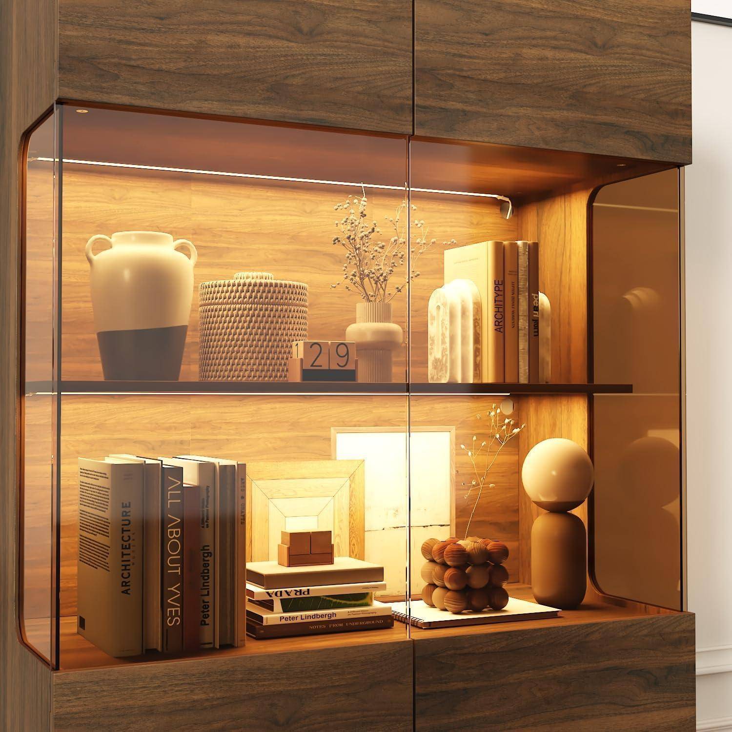 Holzvitrinen­schrank mit warmer eingebauter Beleuchtung, auf zwei Regalen mit Büchern, Vasen, Dekorationsobjekten und einem Bilderrahmen.