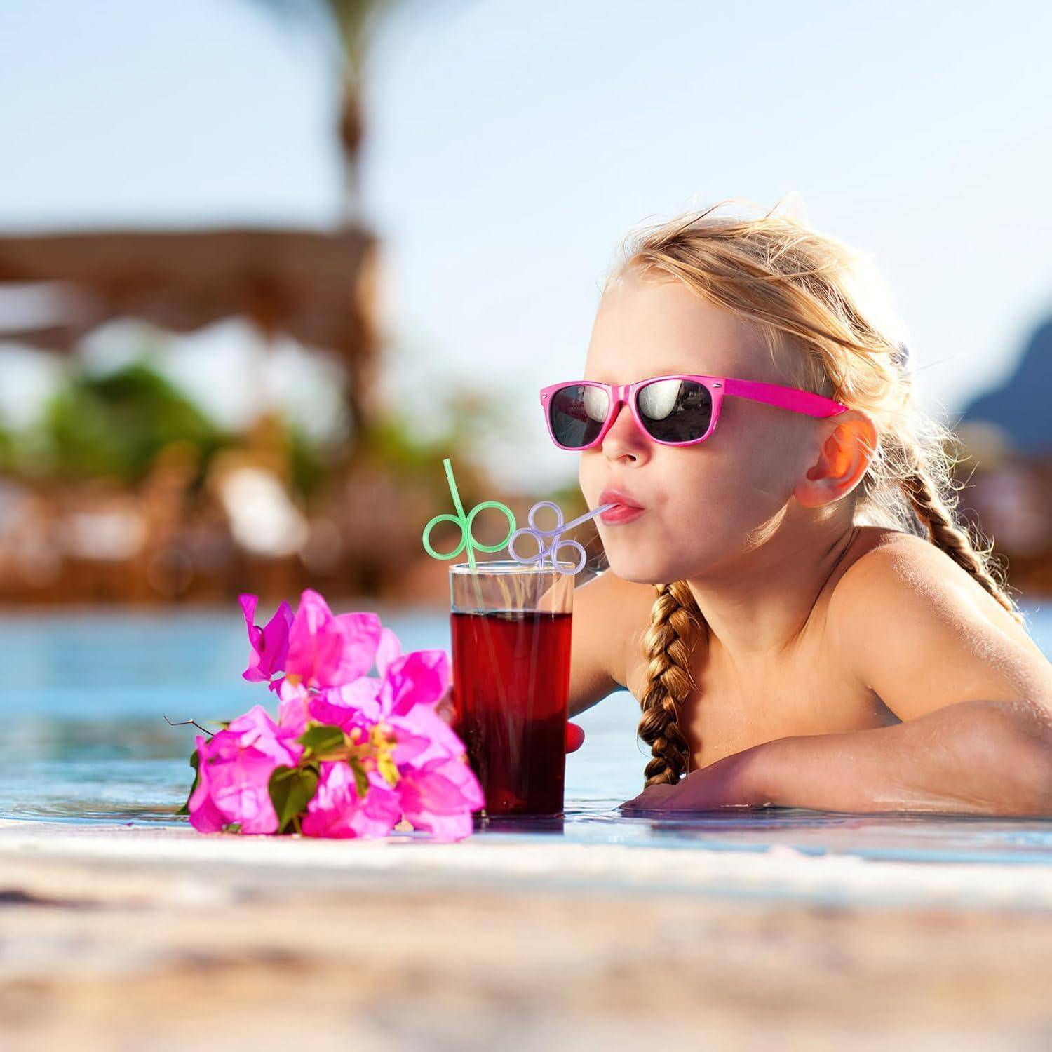 Ein junges Mädchen in rosa Sonnenbrillen entspannt sich in einem Swimmingpool und schlürft ein rotes Getränk mit einem Strohhalm neben rosa Blumen.