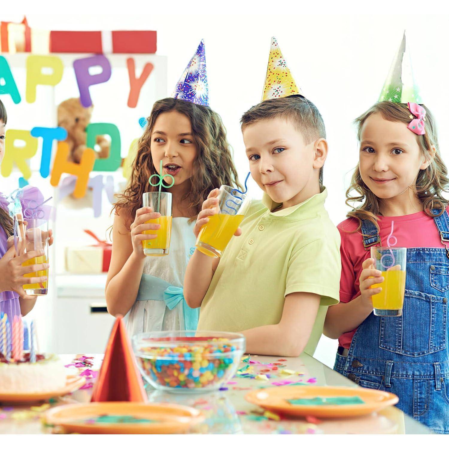 Drei Kinder mit Partyhüten lächeln und halten orangefarbene Getränke an einem Geburtstagtisch, auf dem sich Kuchen und Snacks befinden, mit einem "Alles Gute zum Geburtstag""-Banner im Hintergrund.