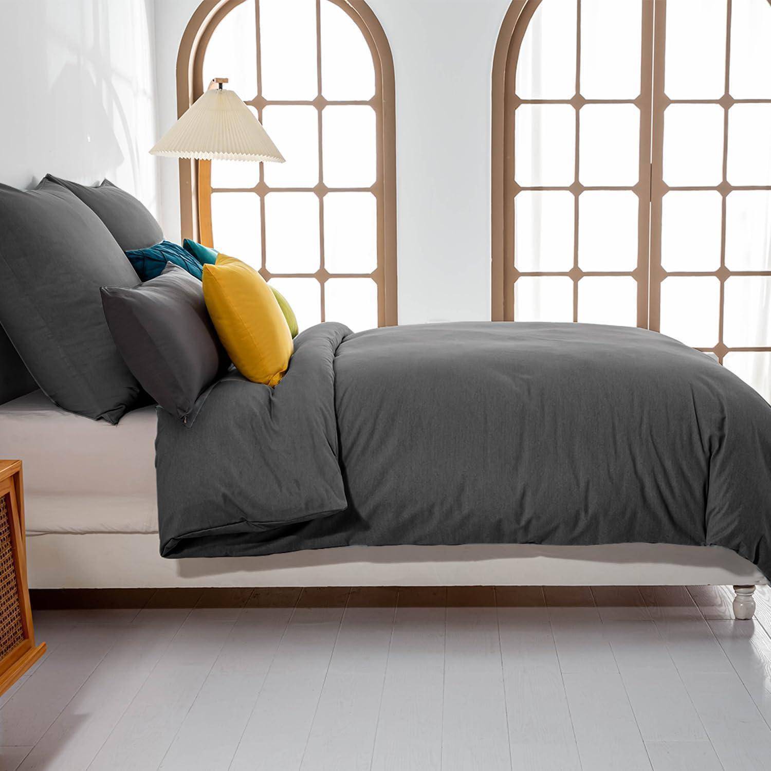 Ein ordentlich gemachtes Bett mit dunkelgrauer Bettwäsche, gelbem Akzentkissen und cremefarbener Lampe in einem hellen Zimmer mit Bogenfenstern.