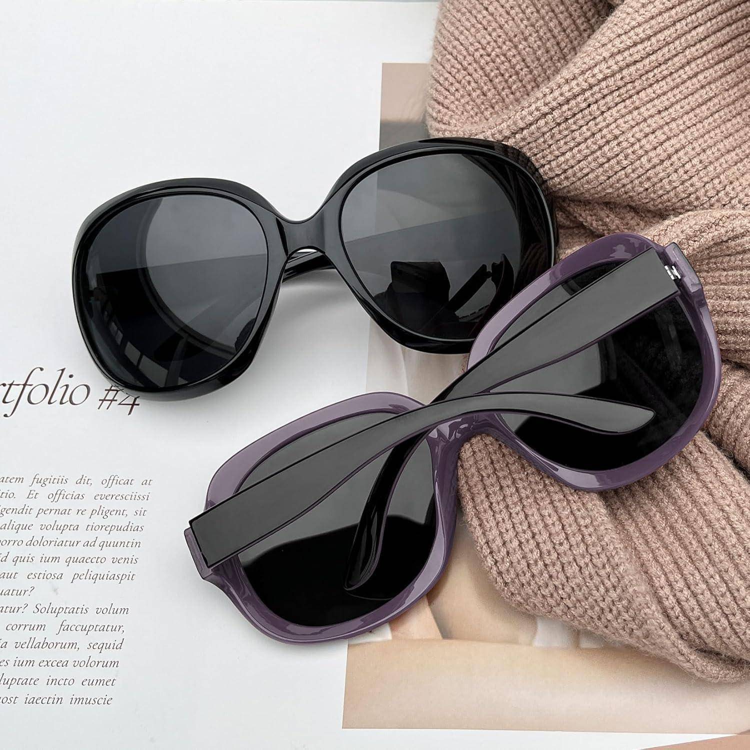 Zwei Paar übergroße Sonnenbrillen, eine schwarze und eine violette, liegen auf einem Magazin neben einem beigen Strickpullover.