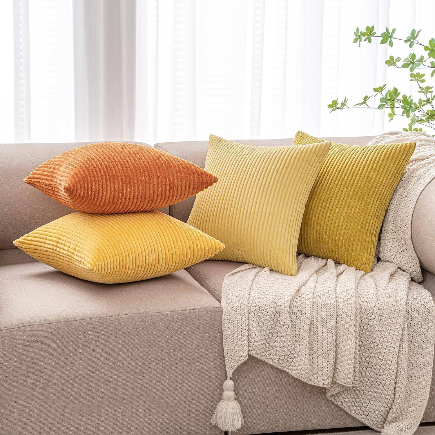 Gestapelte gerippte orange und gelbe Wurfkissen auf einem beigen Sofa mit einer cremefarbenen Strickdecke und weißen Vorhängen.